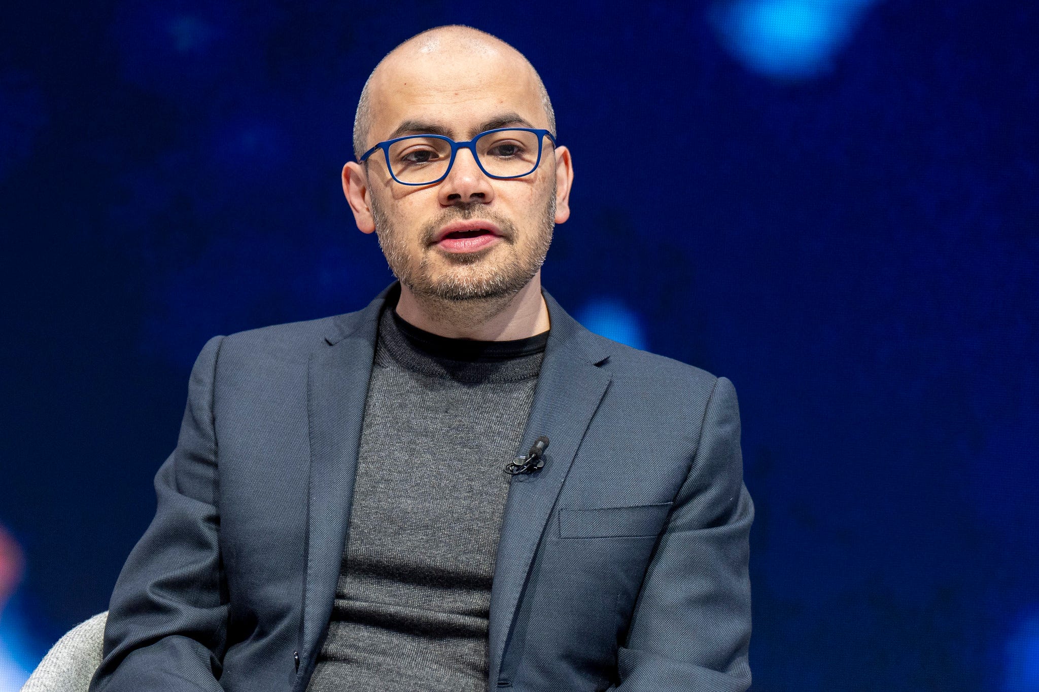 A photo of Google Deepmind CEO Demis Hassabis in a blazer and sweater in front of a blue background