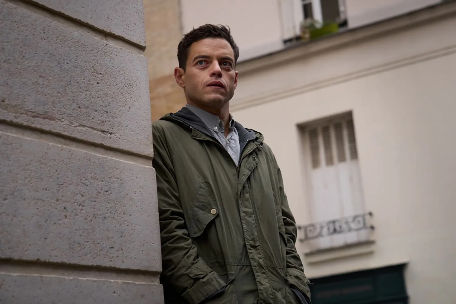Rami Malek in a green coat leans against a concrete wall.