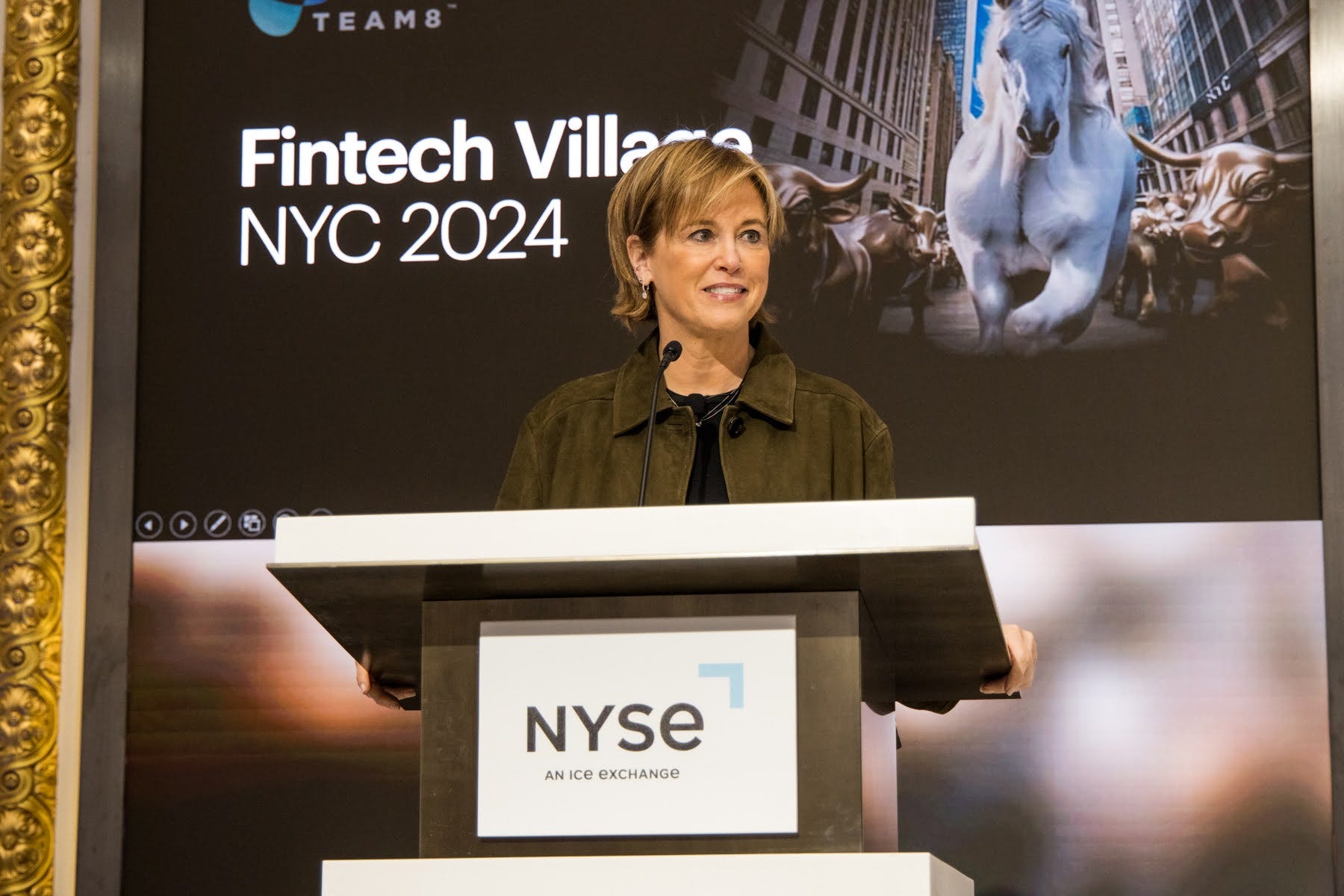 A woman stands at a dais with NYSE on the front. She wears a brown suede jacket and a black shirt. Behind her is a sign that says "Fintech Village NYC 2024"