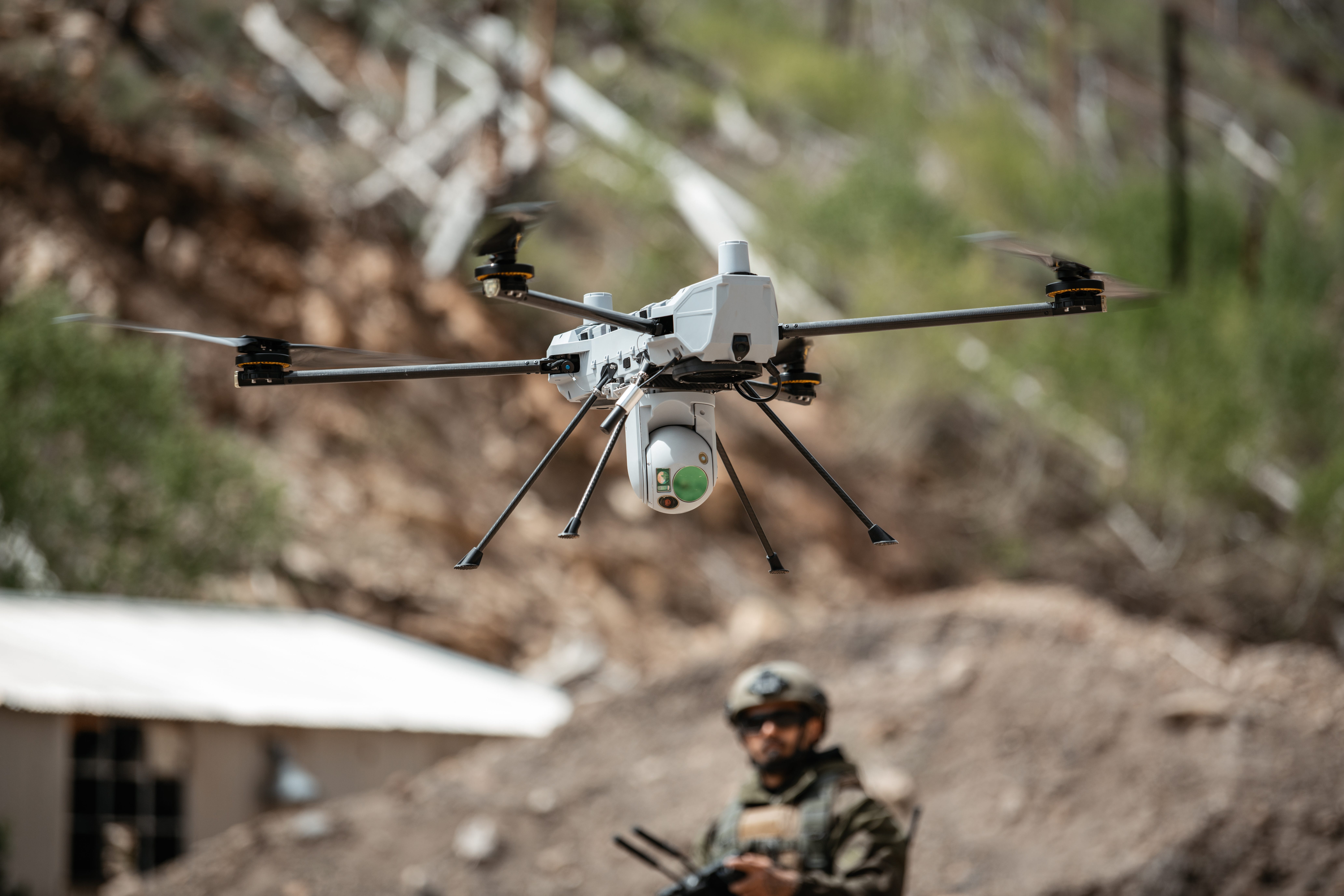 A PDW C100 drone is seen in the foreground with a man wearing camouflage operating it in the background.