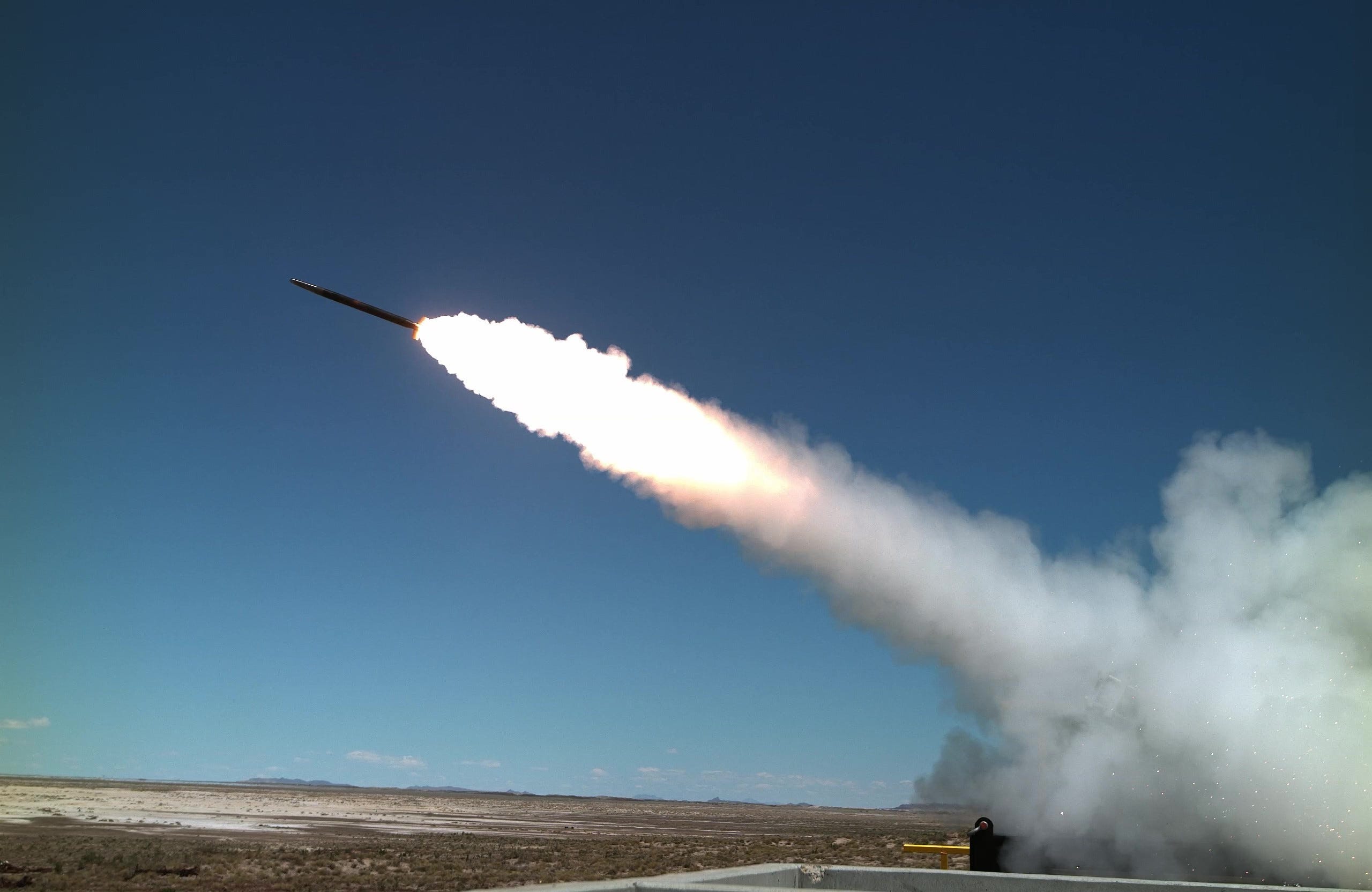 A Stinger missile flies towards the left with a large trail of smoke behind it. The sky is blue in the background and a dusty, barren ground is seen on the bottom.