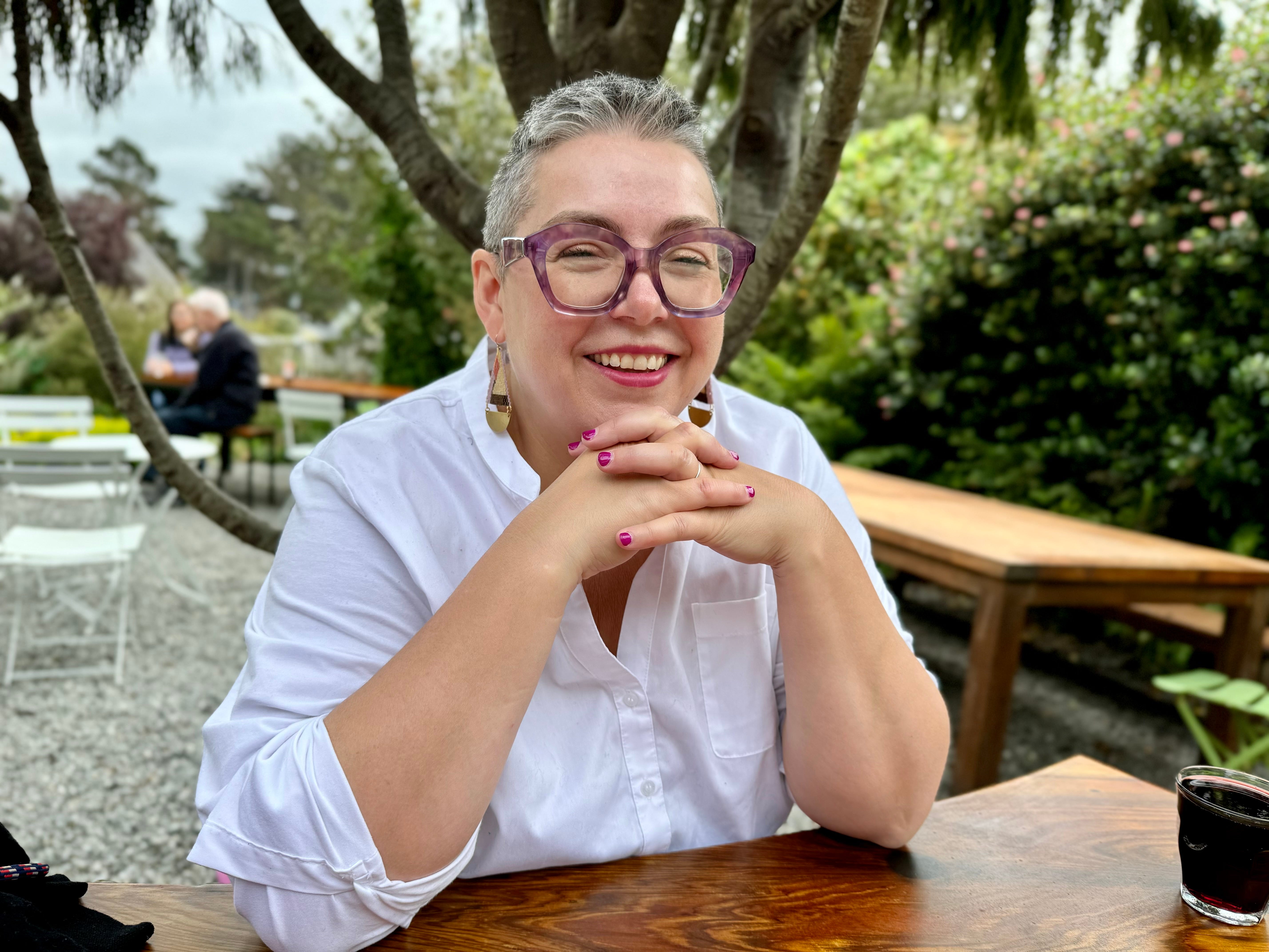 The author wearing purple glasses and looking at the camera while smiling and sitting outside at a wooden table.
