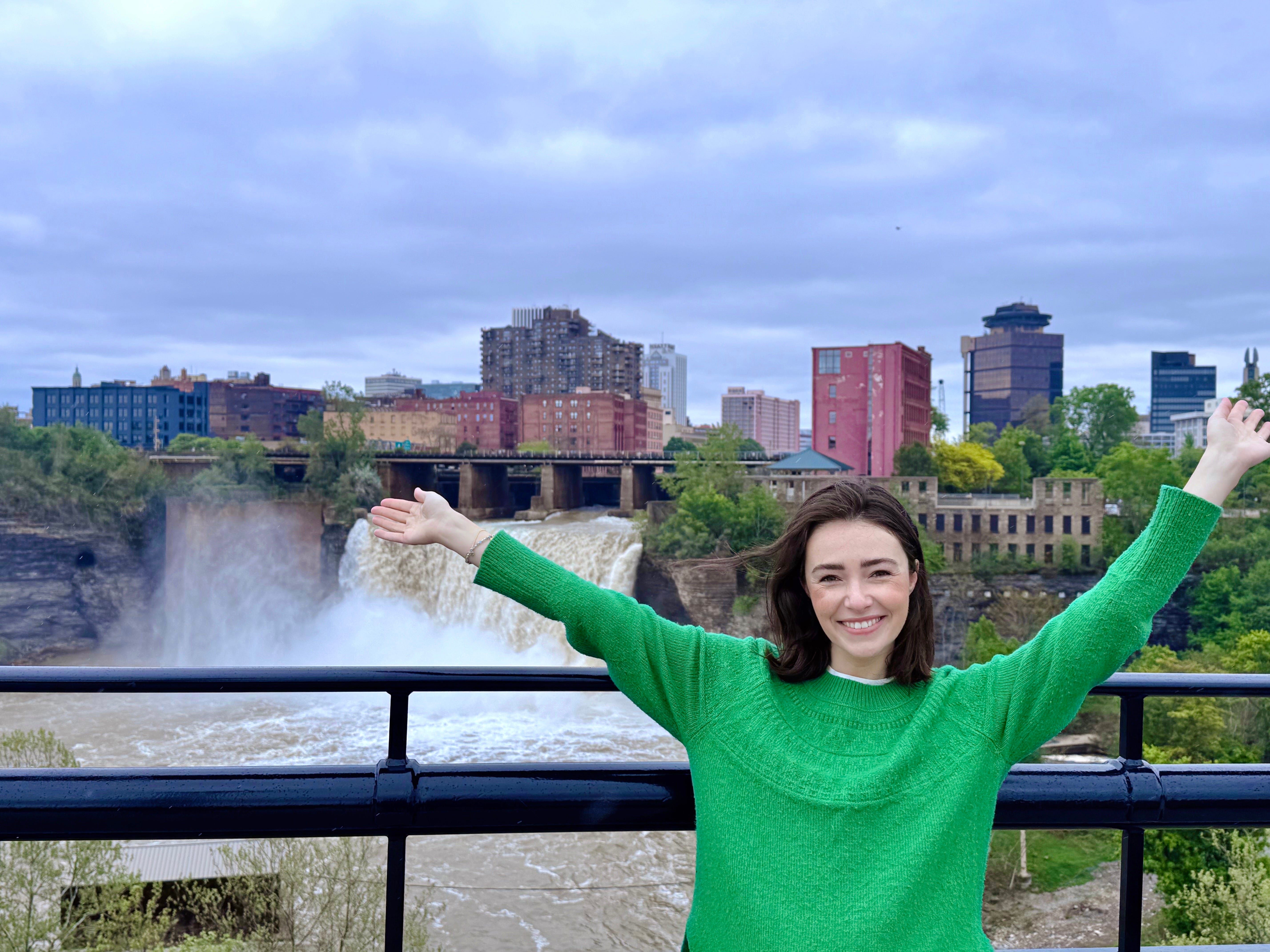 The author smiling and posing with her arms up at the High Falls in Rochester, New York. She's wearing a green long-sleeved sweater.