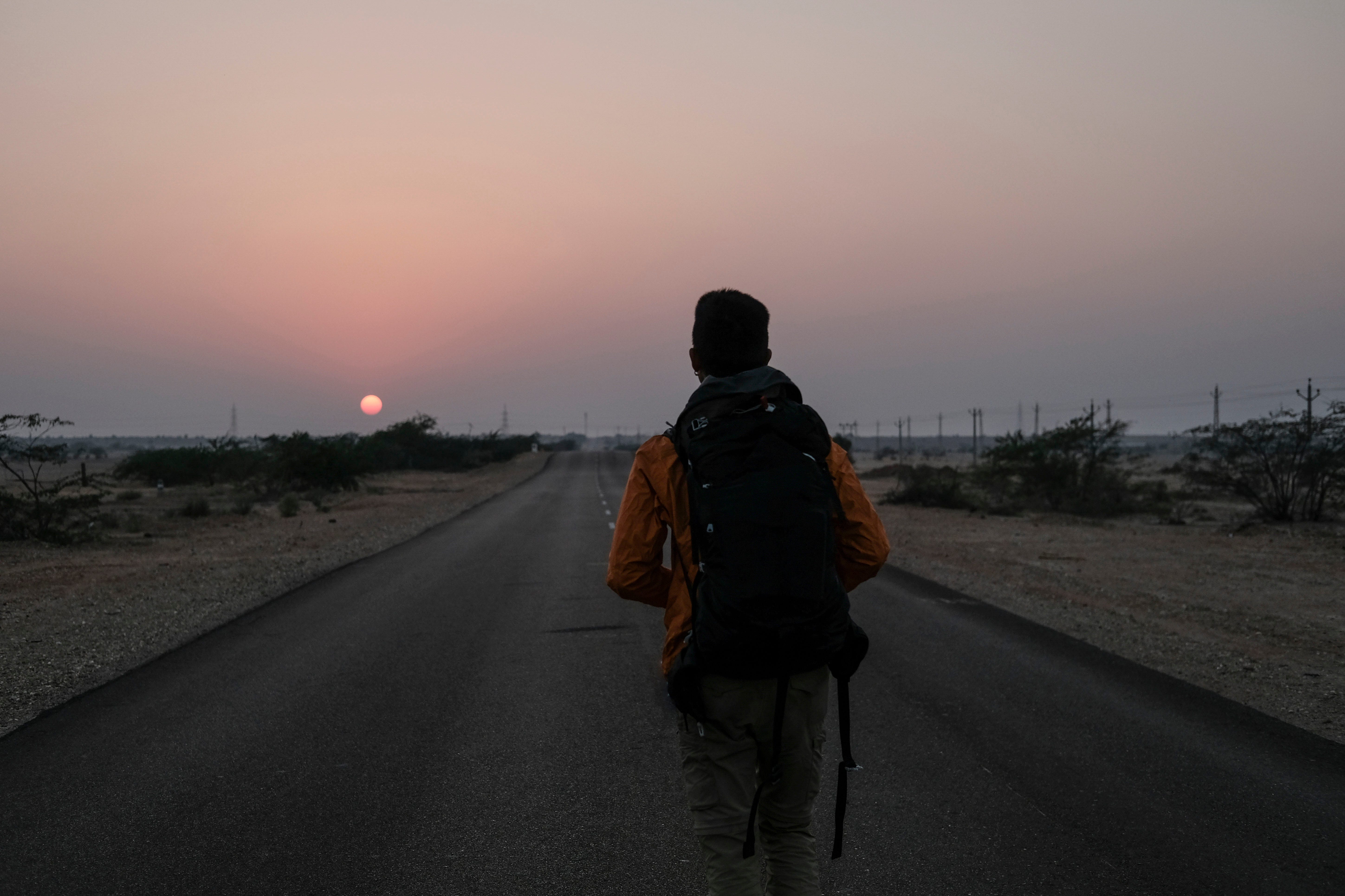 Man with backpack walking on country road against sky during sunset