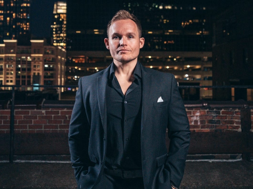 A clean-shaven man with short blond hair stares at the camera. He's wearing a black suit jacket and a black shirt with an open collar. There is a silver handkerchief in his left breast pocket. He's stood on a rooftop and there is a brick wall behind him. There are skyscrapers in the background that are lit up against the night sky.