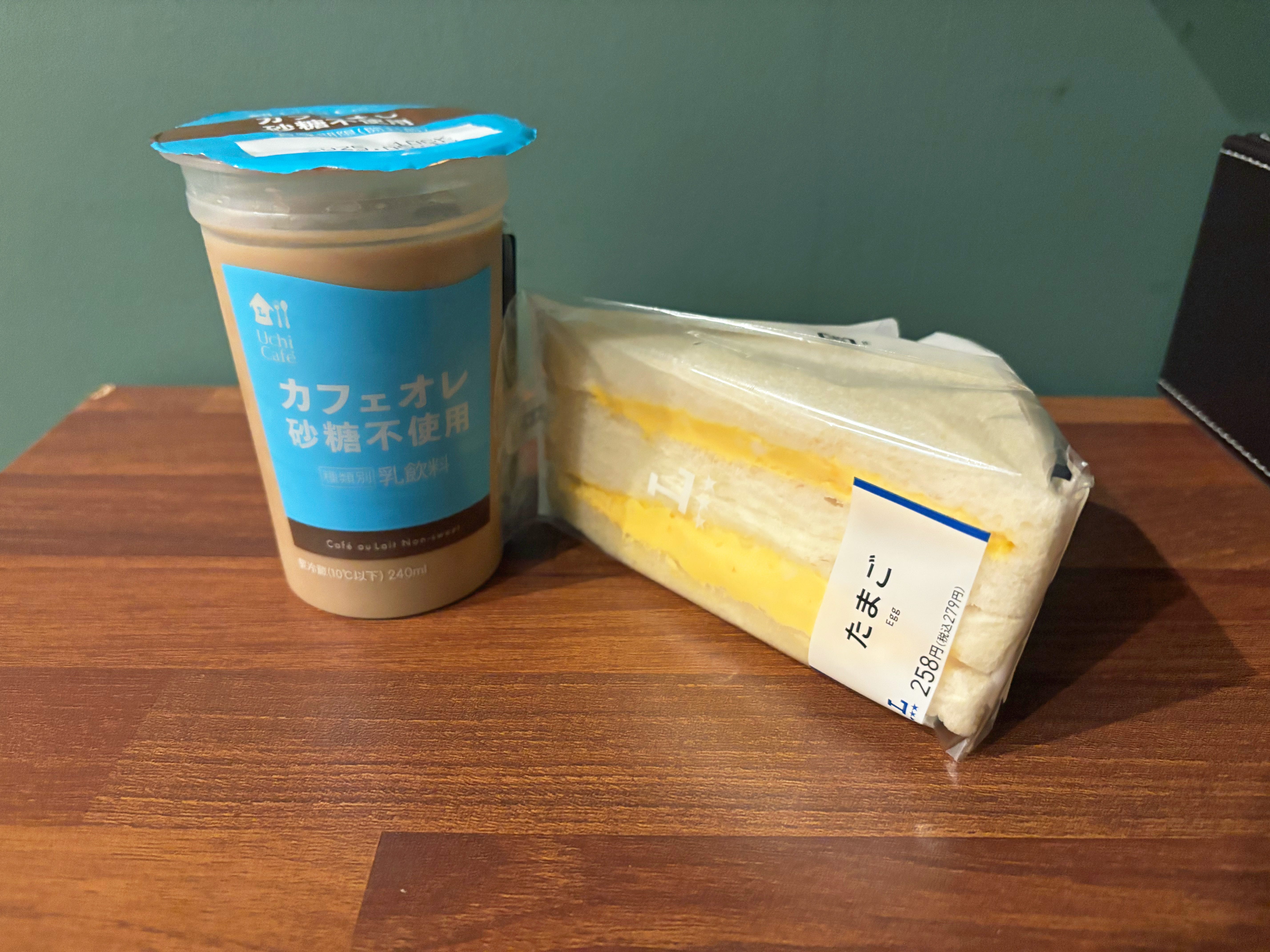 A cup of coffee with Japanese writing next a plastic-wrapped sandwich on a wooden desk