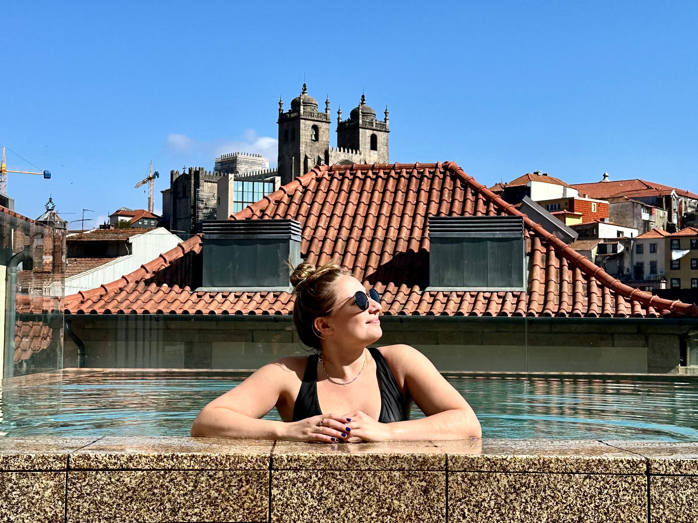The author sits in a rooftop pool, wearing a black bathing suit and sunglasses. Behind her are the tops of buildings and a church.