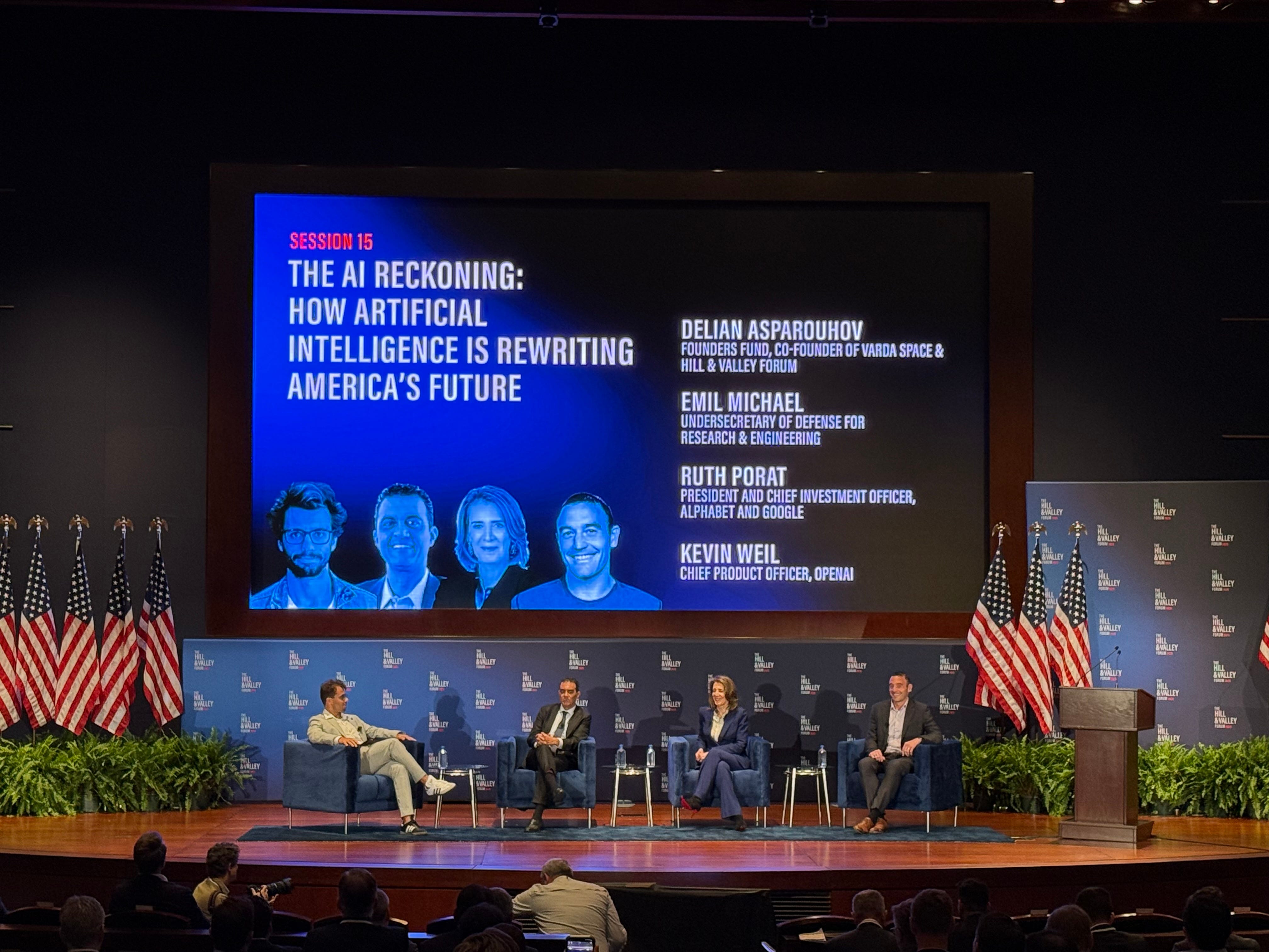 A panel of four Hill and Valley speakers on stage, flanked by American flags.