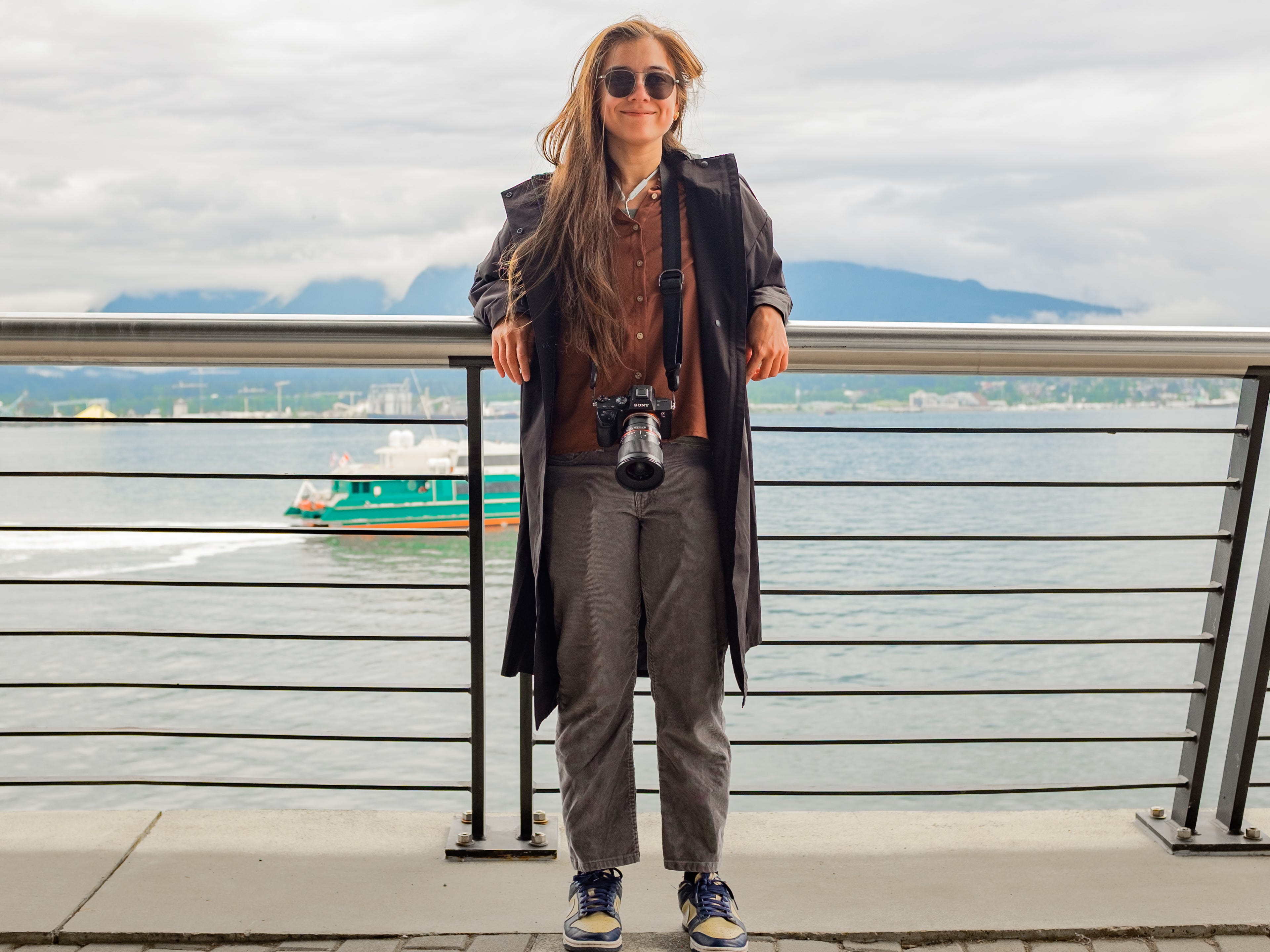 The author leans against a railing in front of the ocean with mountains behind on a cloudy day in Vancouver