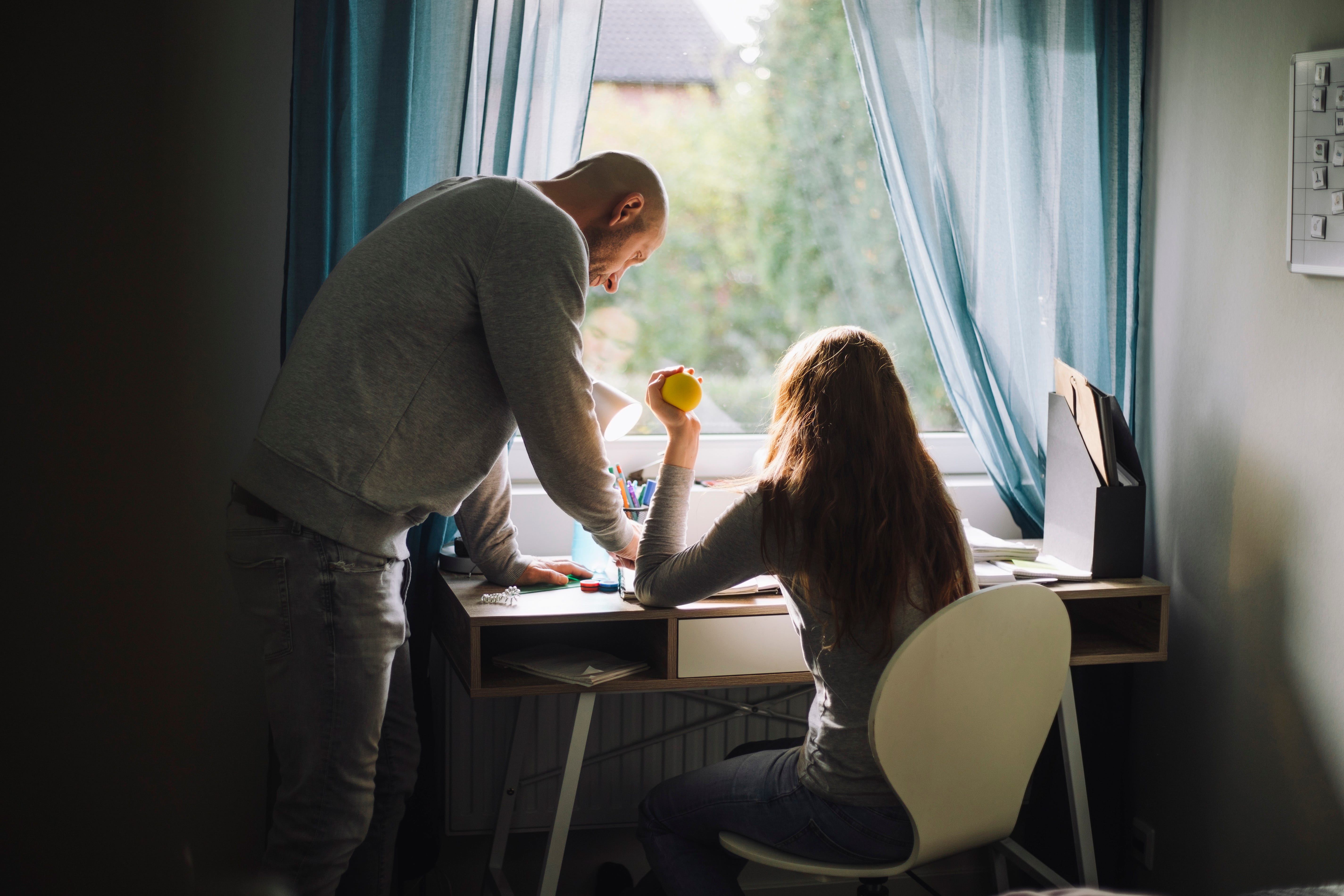 A girl sits on a desk, as a man looks down on the desk.