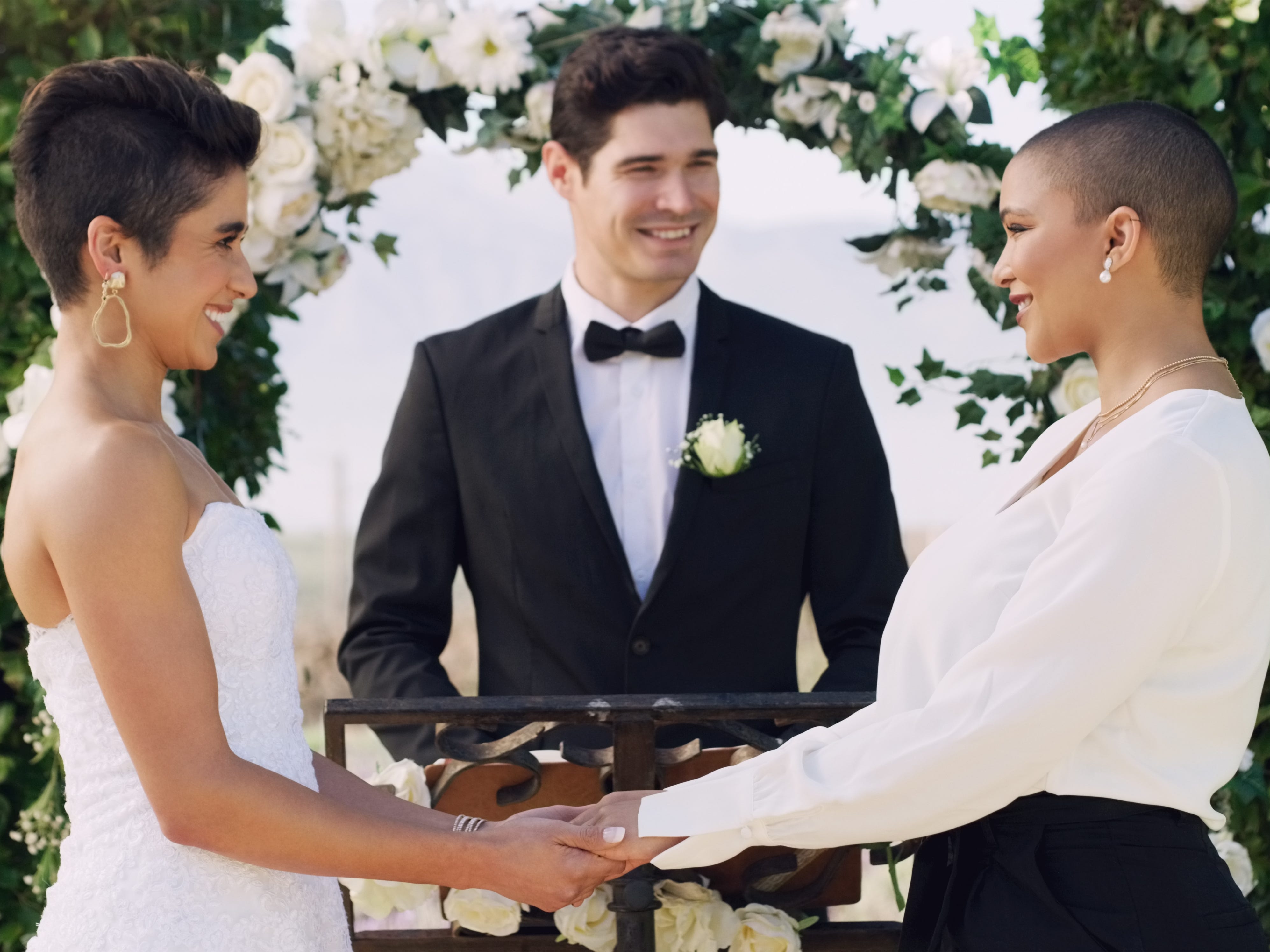 Two brides hold hands at their wedding ceremony. Their officiant smiles in the background.