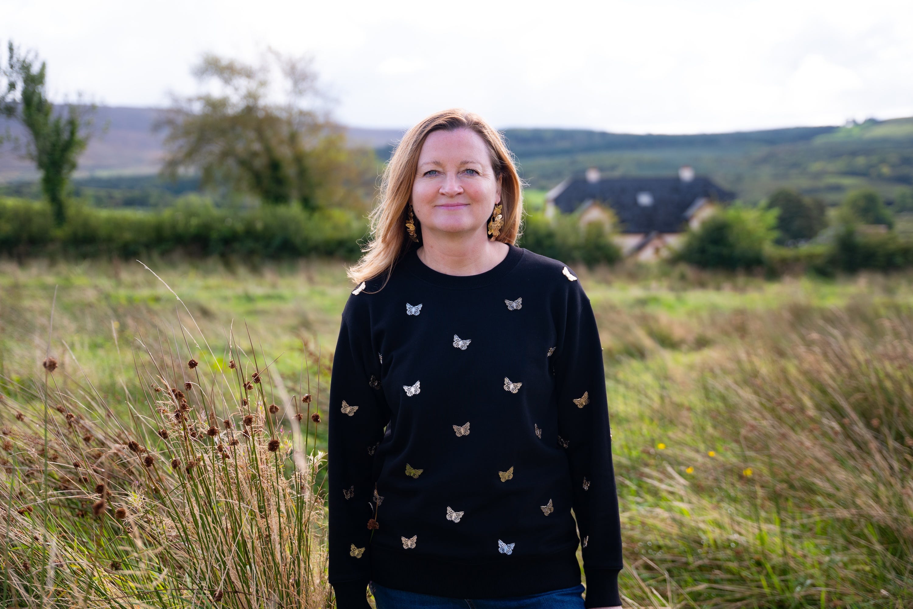 A woman in a black sweater with metallic butterflies it and butterfly earrings stands amid tall grass with rolling hills in the background