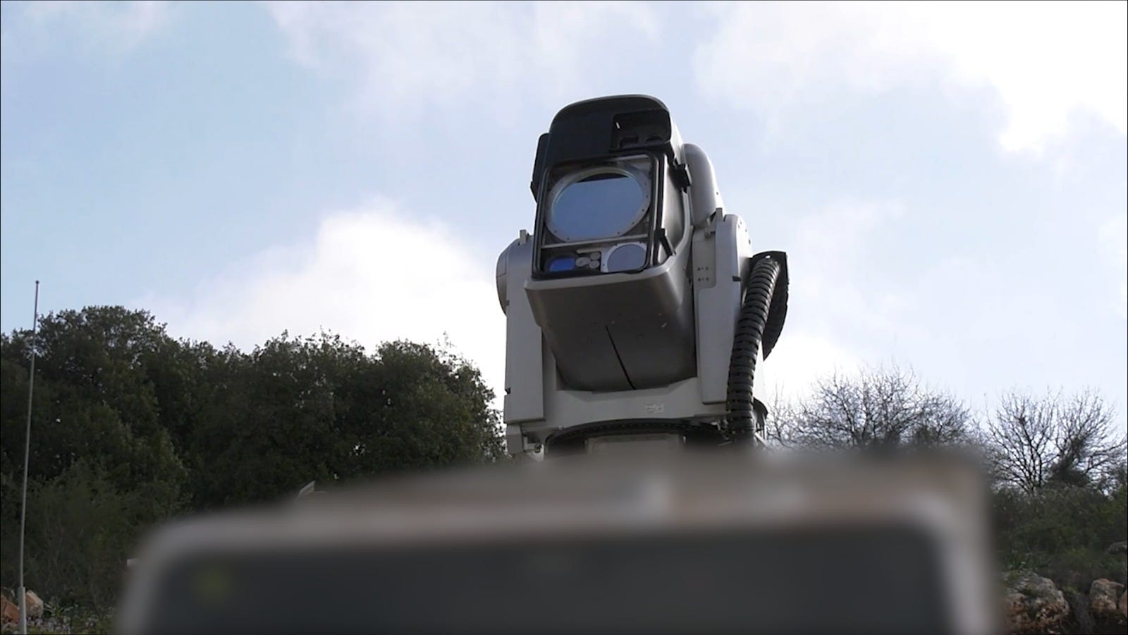 A close-up low angle of Israel's laser weapon with a line of trees and a cloudy blue sky in the background.