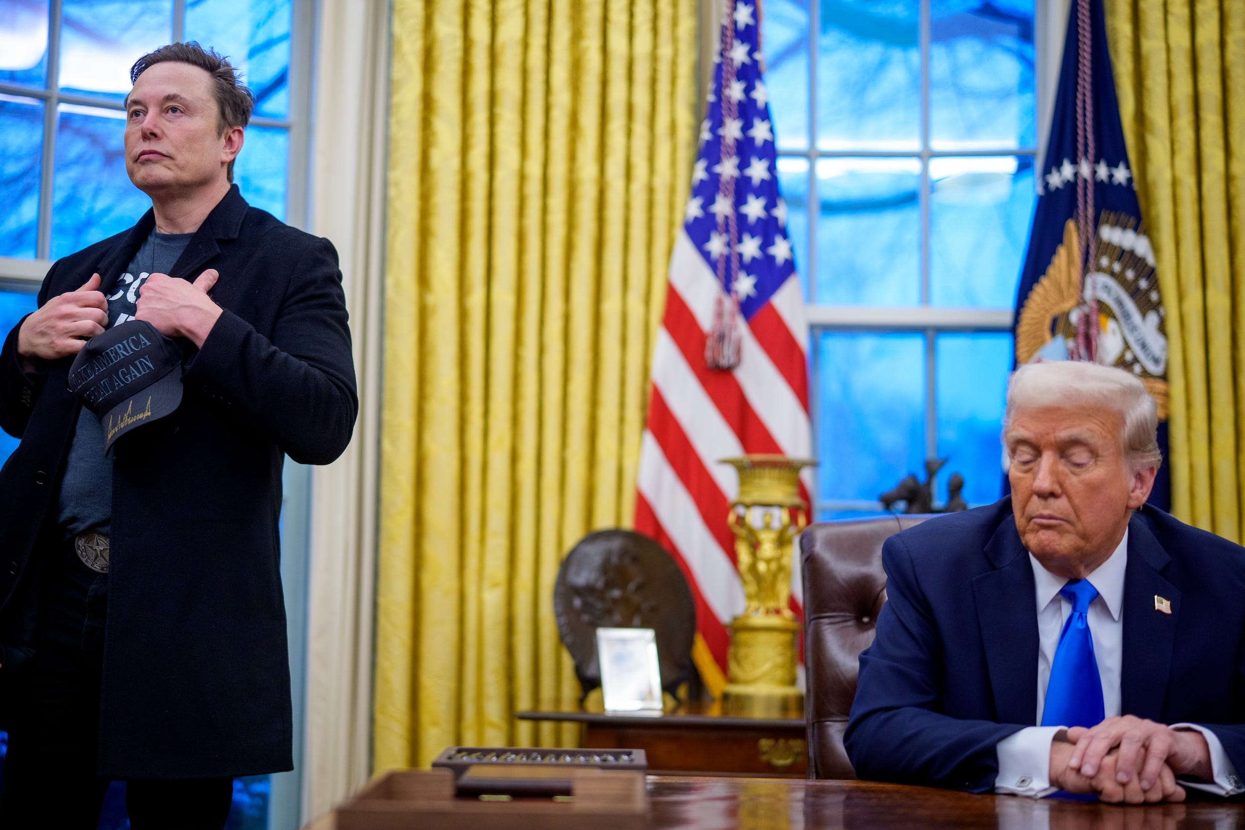 Elon Musk looks out at reporters as Donald Trump looks down at the Resolute Desk in the Oval Office