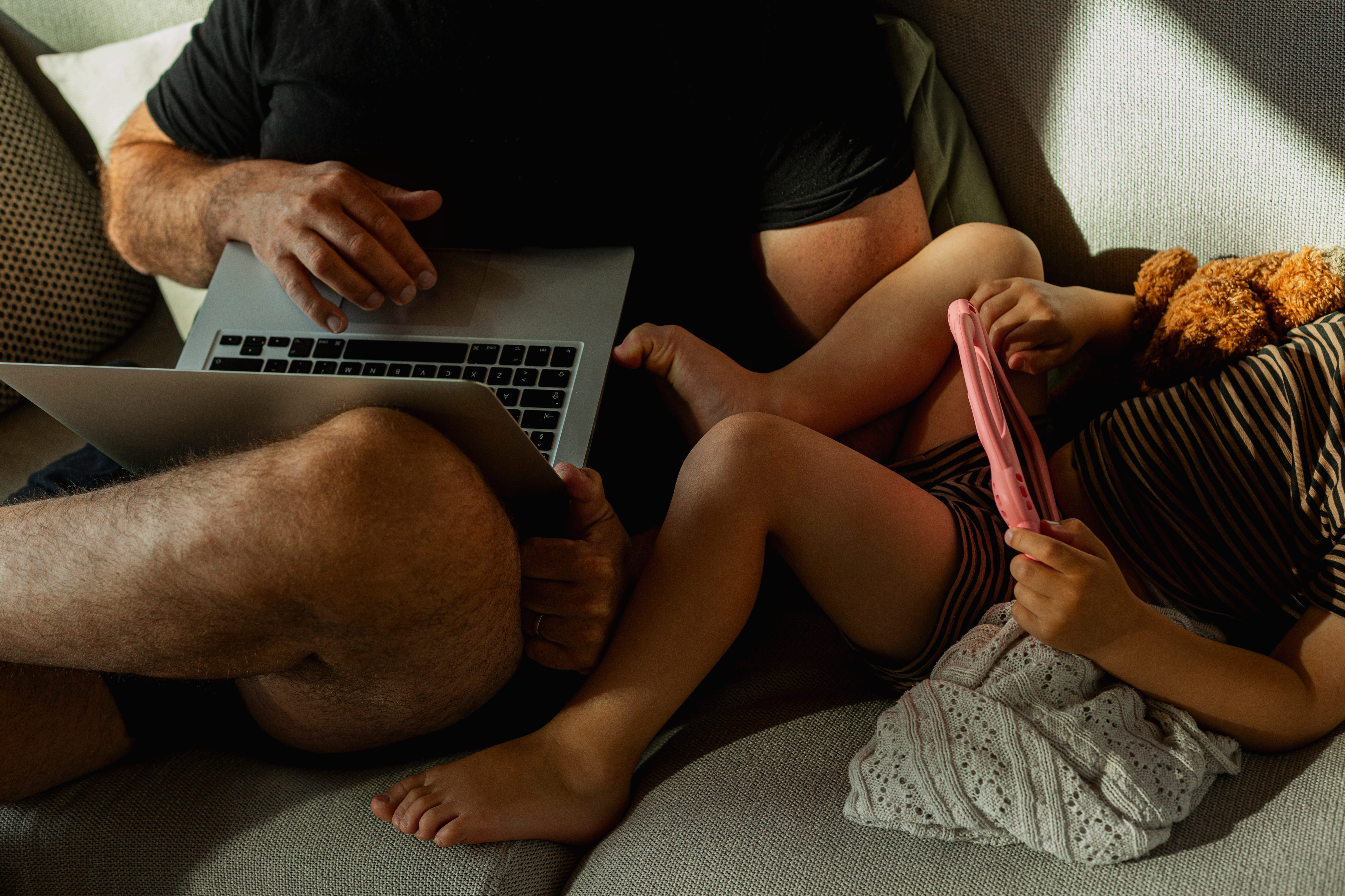 Man tying on computer next to kid with ipad on couch