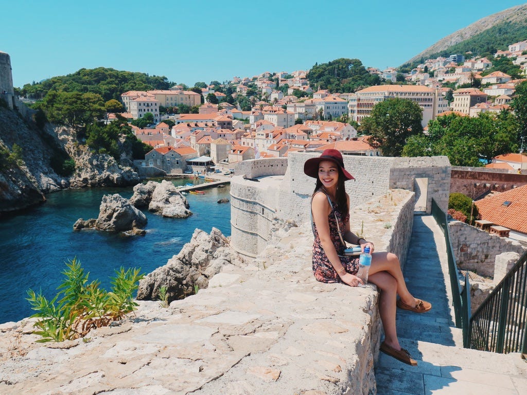 Veena sits on a wall near the water, looking over her shoulder. Behind her, a city with white buildings and orange roofs is visible.