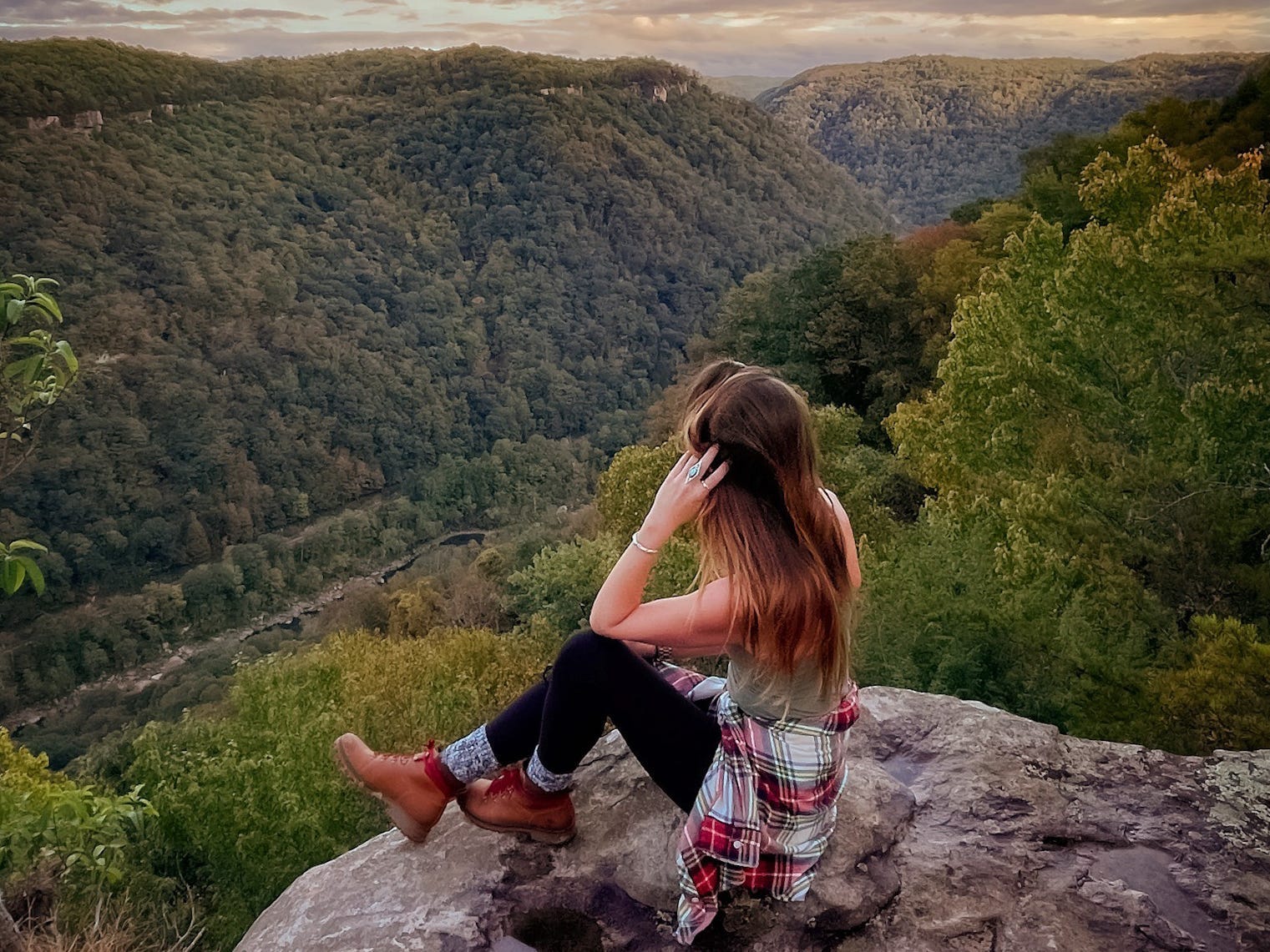 Emily, wearing a green top, a flannel around her waist, black leggings, high socks, and hiking boots, sits on a mountain looking out at the trees.