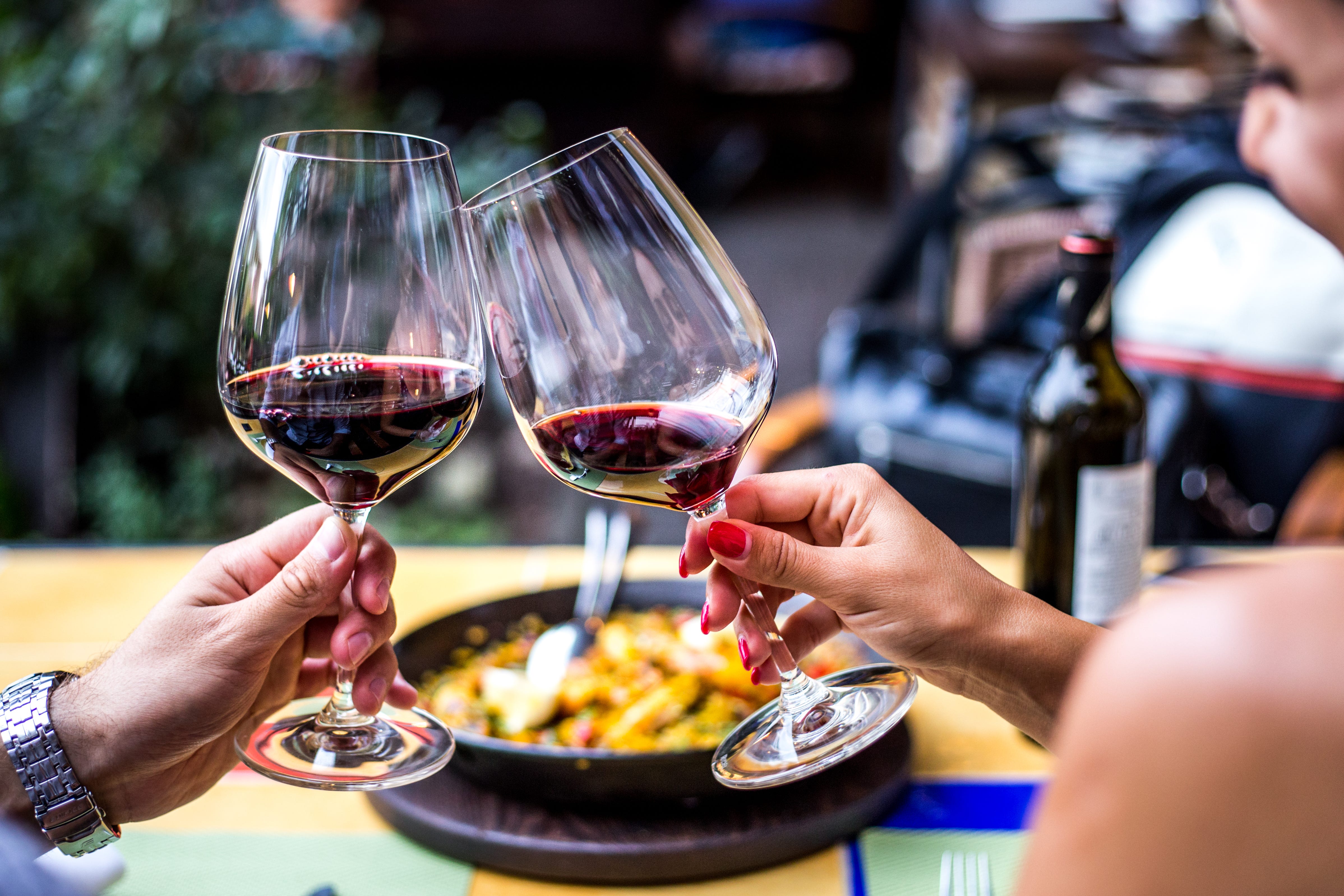 Two people touching wine glasses with red wine together in front of meal
