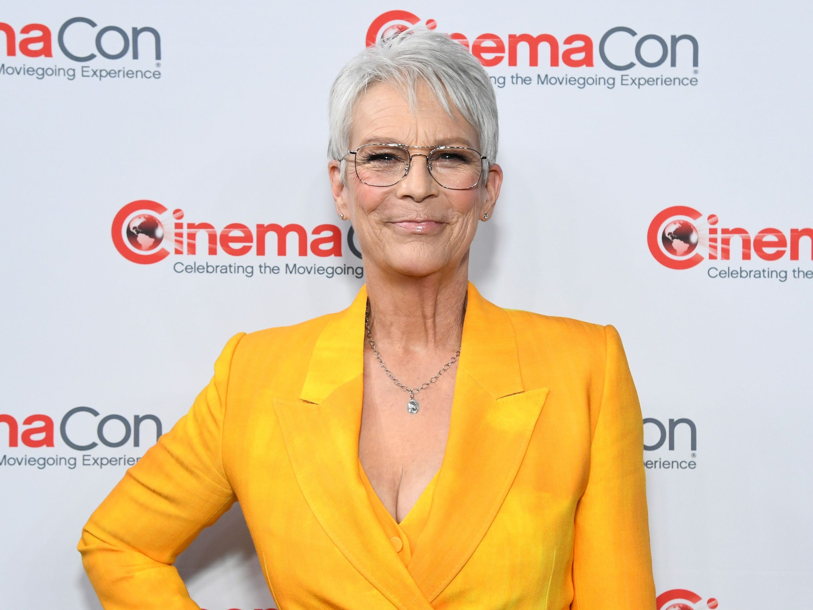 An older woman with short gray hair and large-framed glasses wears a bright yellow suit. She's also wearing a silver necklace. There is a red logo for CinemaCon on the white wall behind her.
