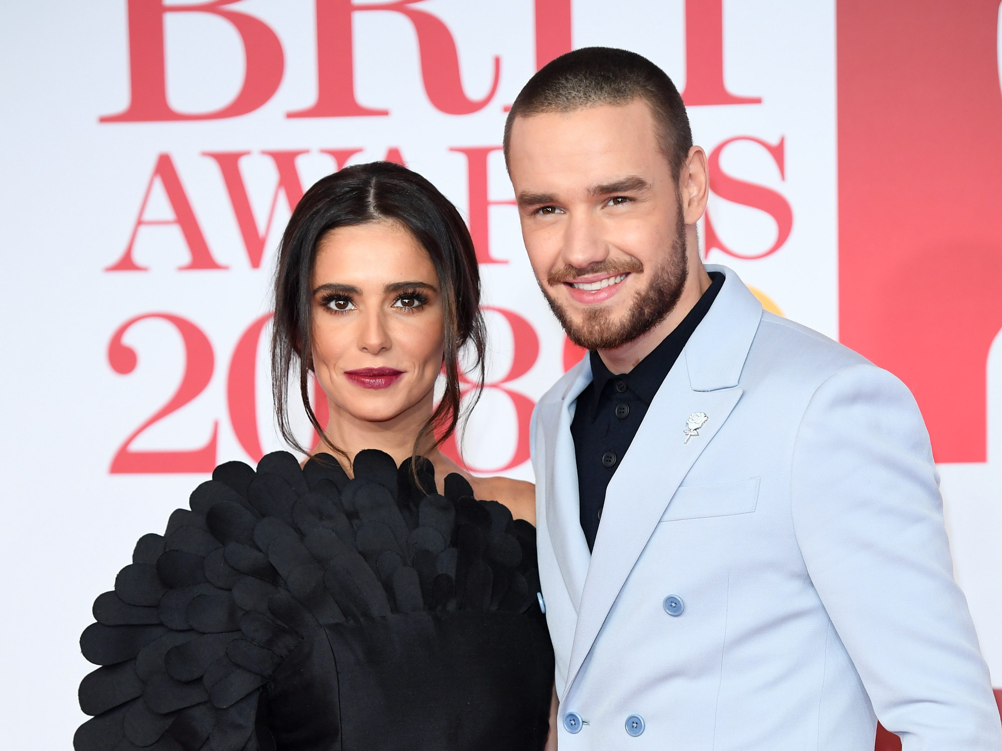 A woman and a man pose on a ride carpet. On the left, the woman has brown hair tied back with long strands of her fringe hanging around her face. She's wearing a black dress with a feathered effect down her right sleeve. ON the right is a man with a short buzzcut, he has a short brown beard. He's wearing a baby blue double-breasted blazer jacket and a black shirt.