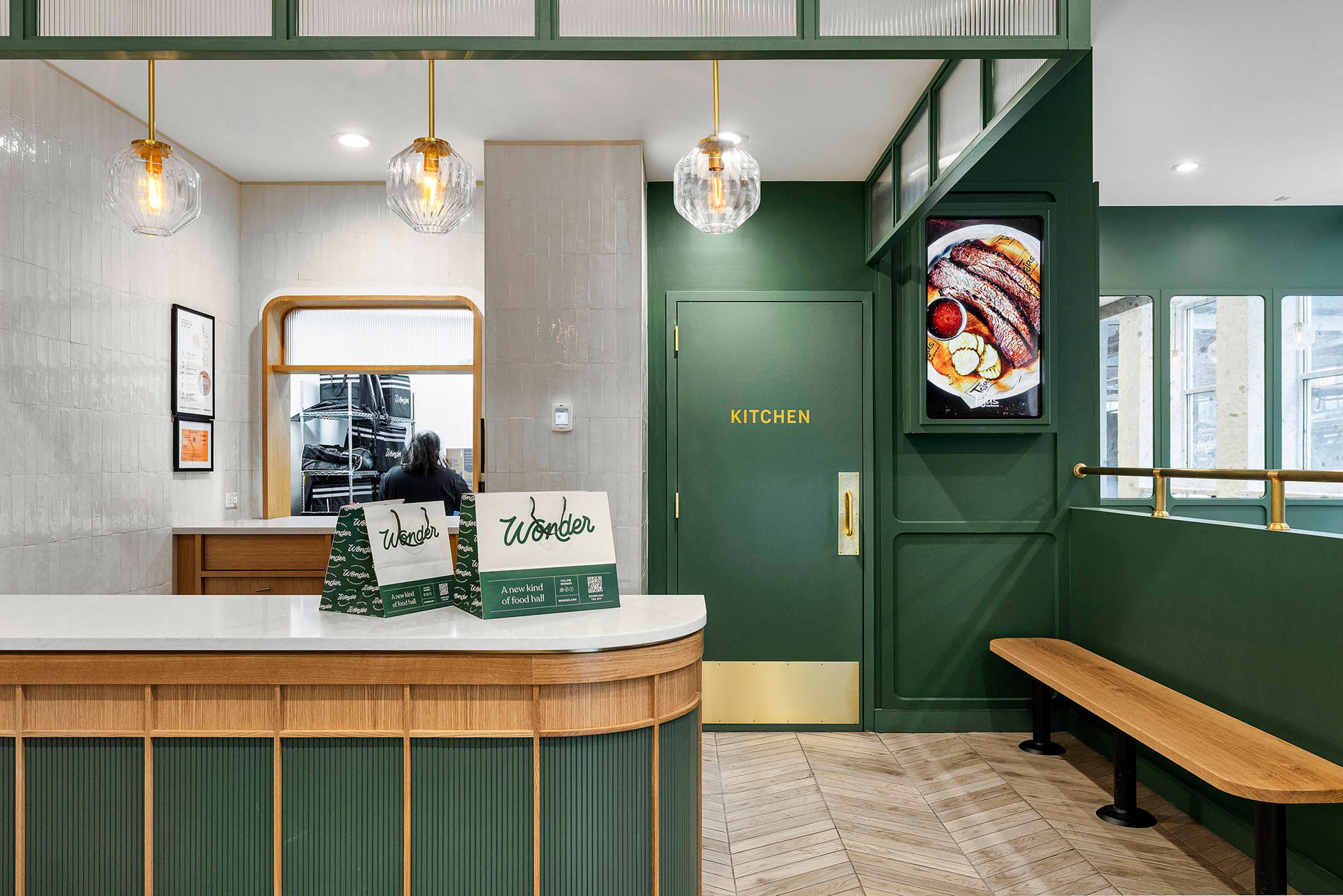 Two paper bags for Wonder food orders sit on the counter of a Wonder restaurant with green and white walls and a door that says "Kitchen" on it in the center. To the left, a worker with their back to the camera looks through a window into the kitchen.