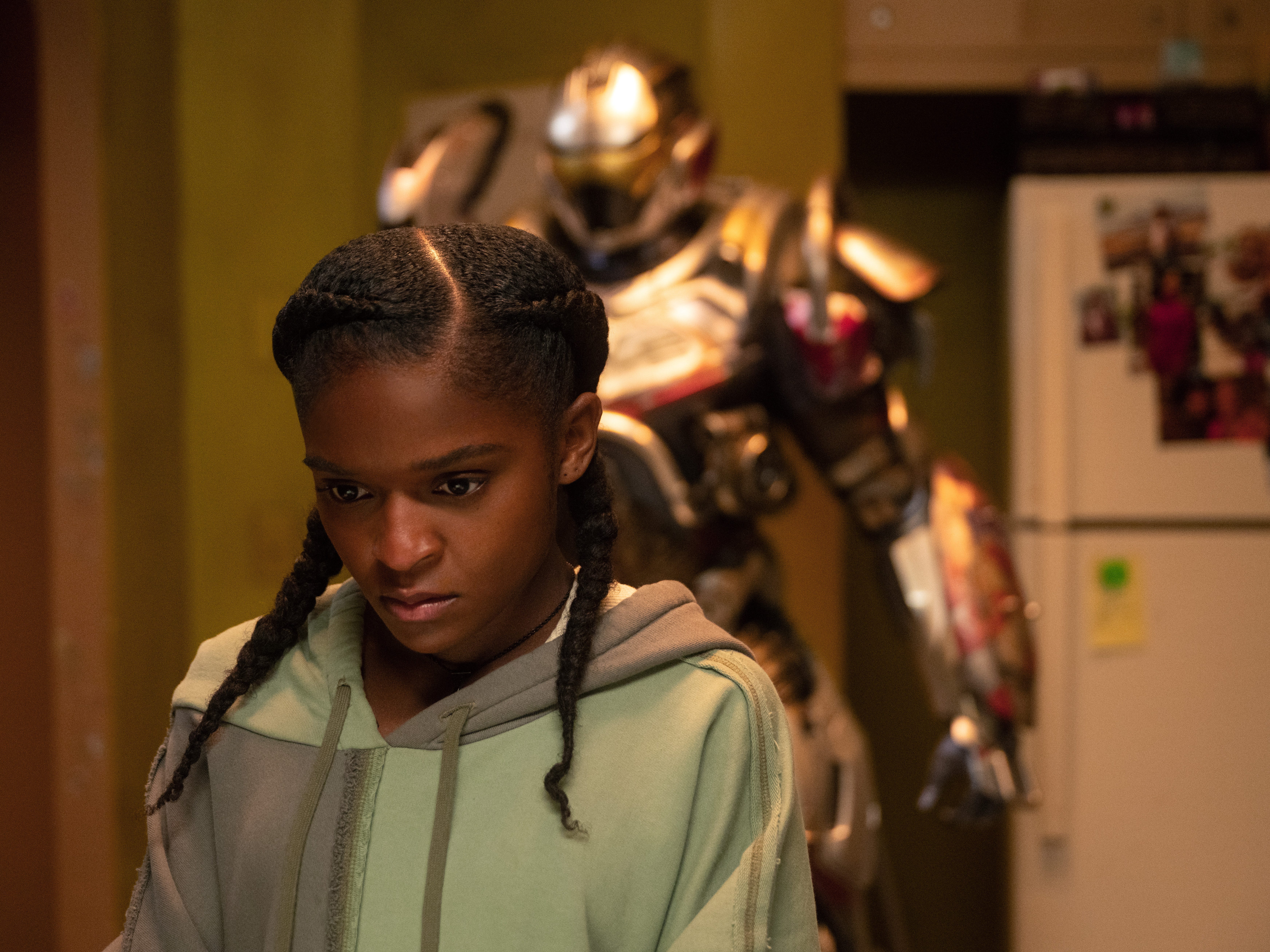 A Black teenage girl with short braids looks down at something. She's wearing a green and gray hoodie. There is a large suit of armor behind her in a kitchen next to a fridge.