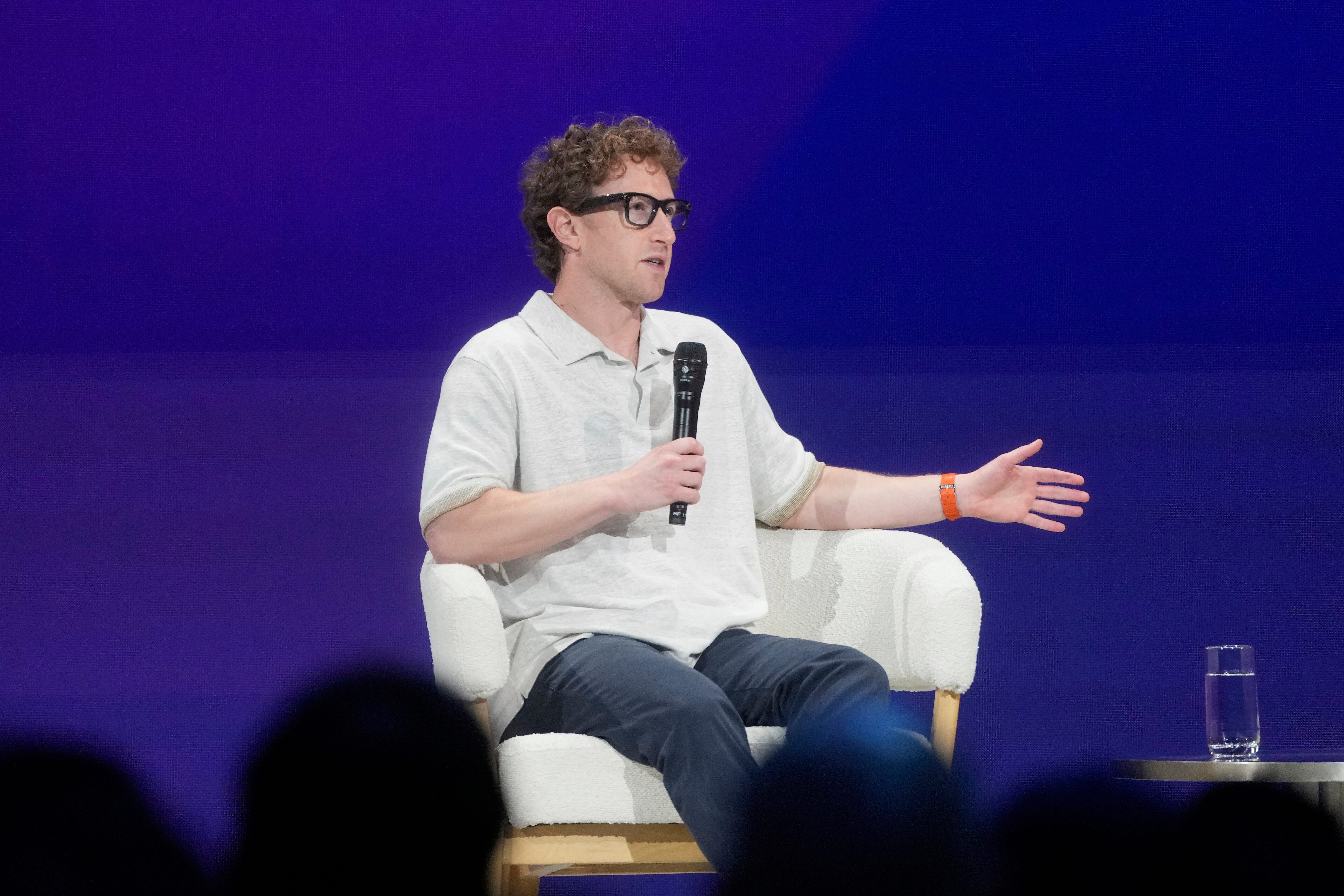 Mark Zuckerberg, a white man in a grey polo shirt and dark pants sits in a white chair holding a microphone in front of a dark purple background.