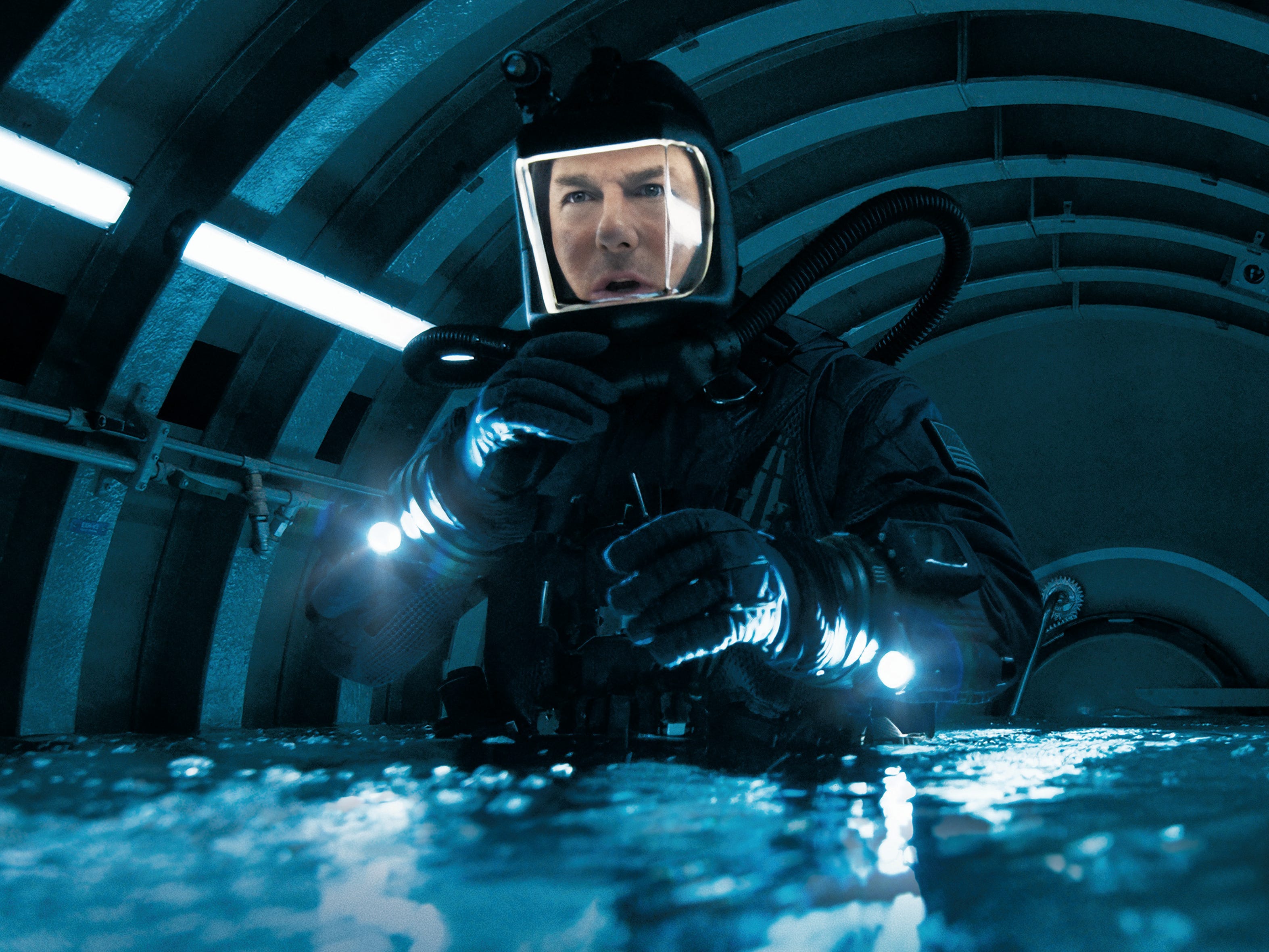 A man in a scuba diving outfit with an illuminated mask is stood in a circular room that is filling up with water.