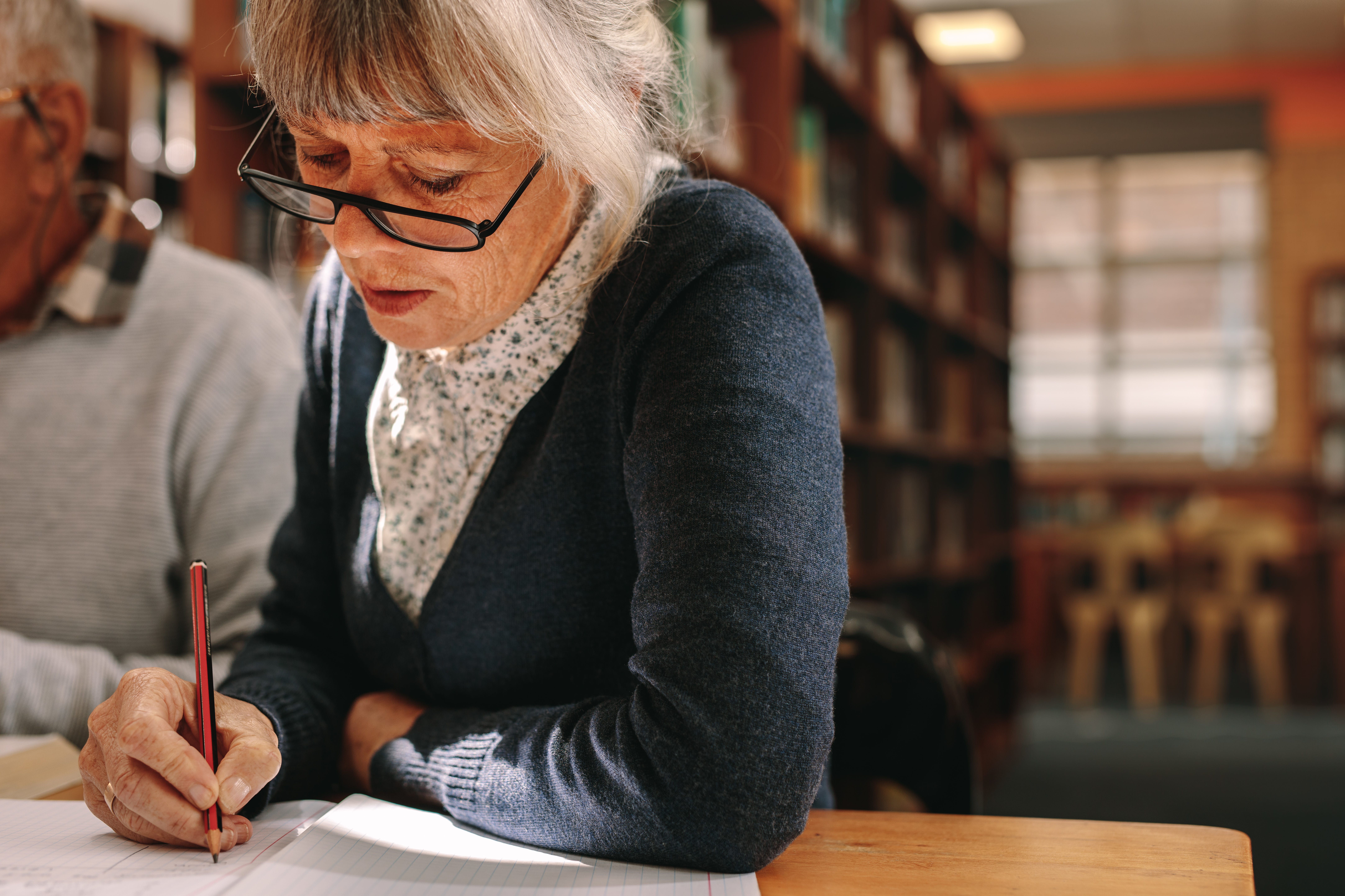 An elderly woman studies in a library setting with her husband.