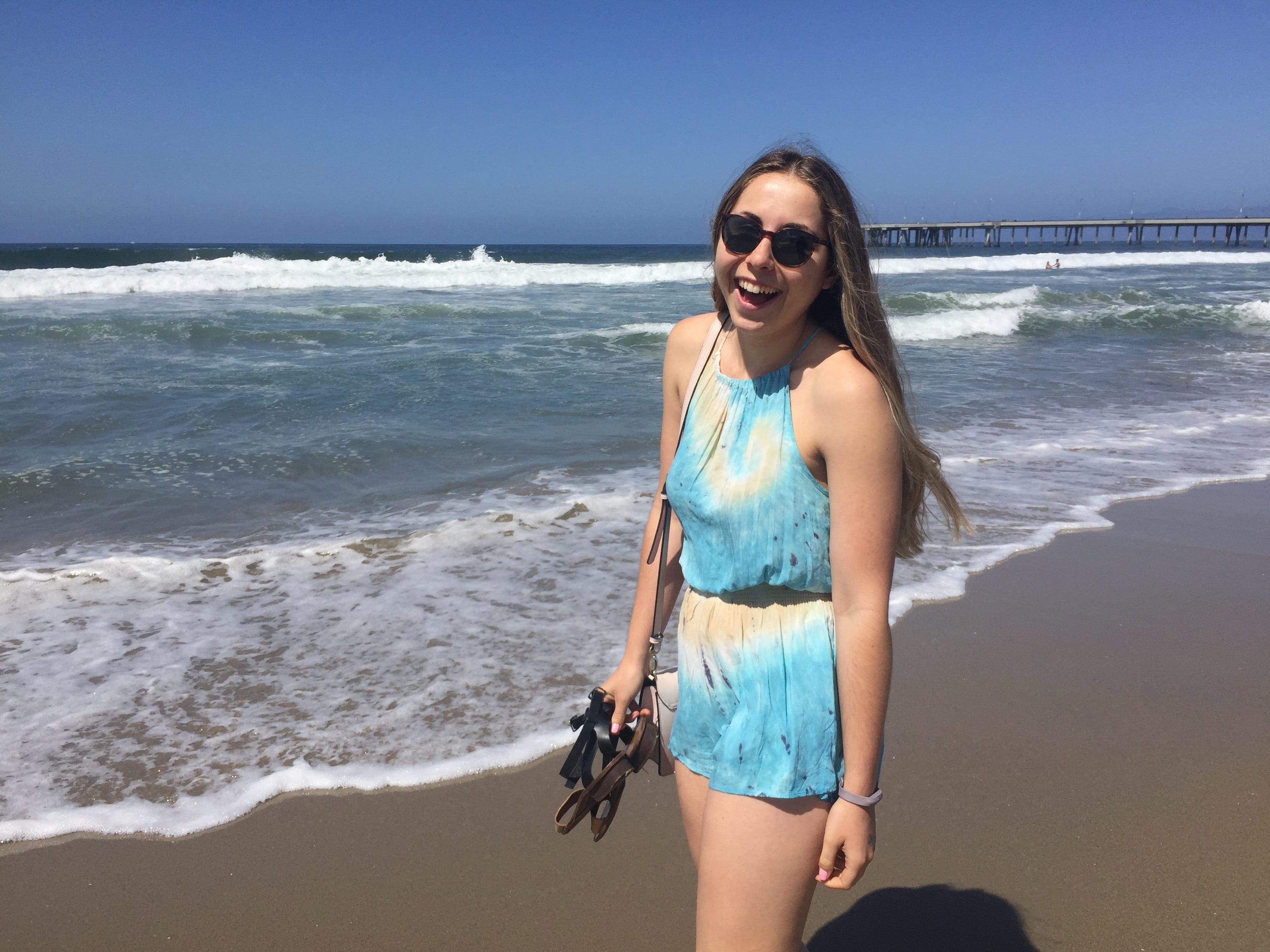 The author wearing sunglasses and a blue and white romper standing on a beach and smiling.