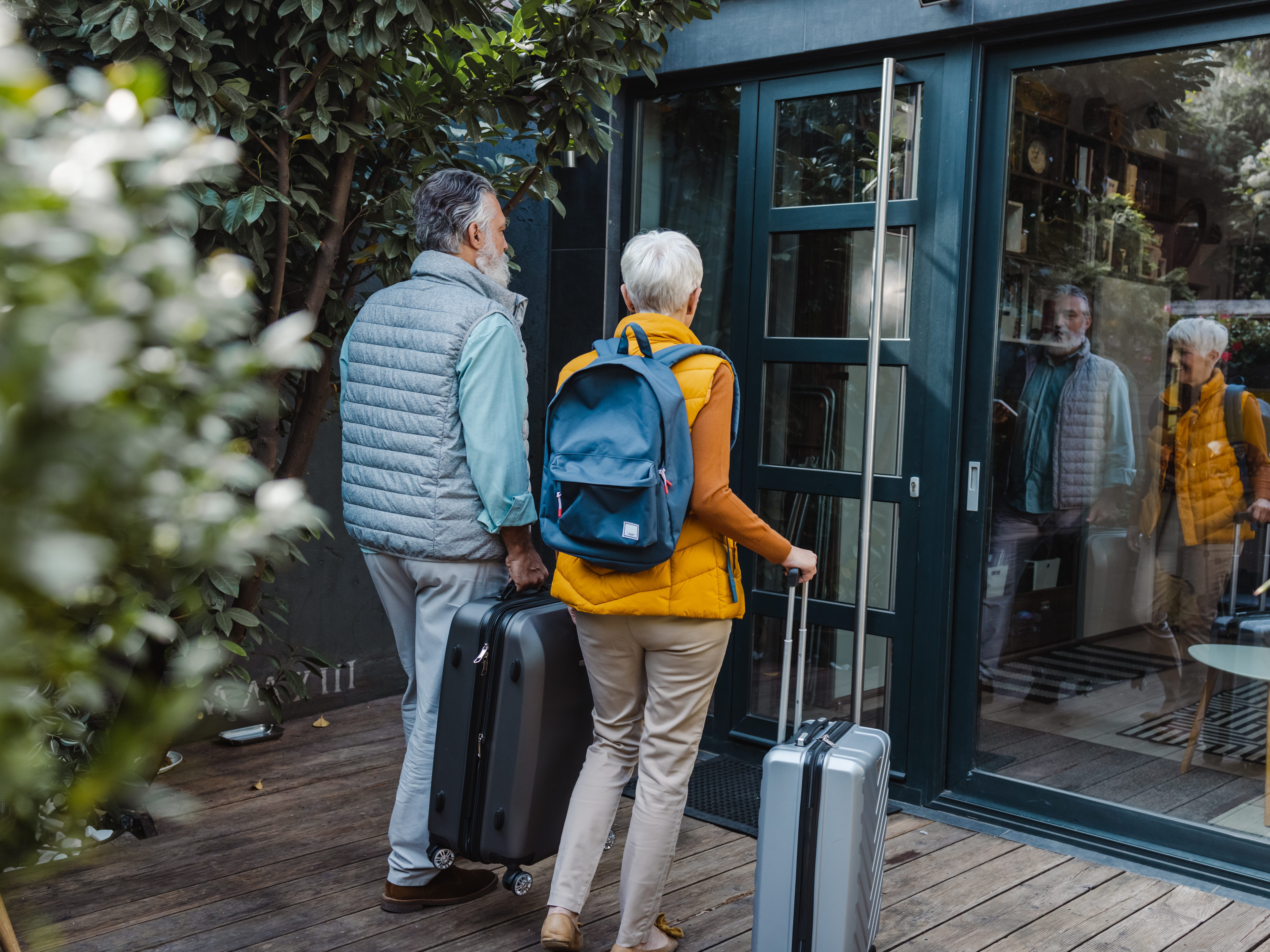 A couple walking into an apartment with suitcases, they are standing on the deck and walking toward the door.