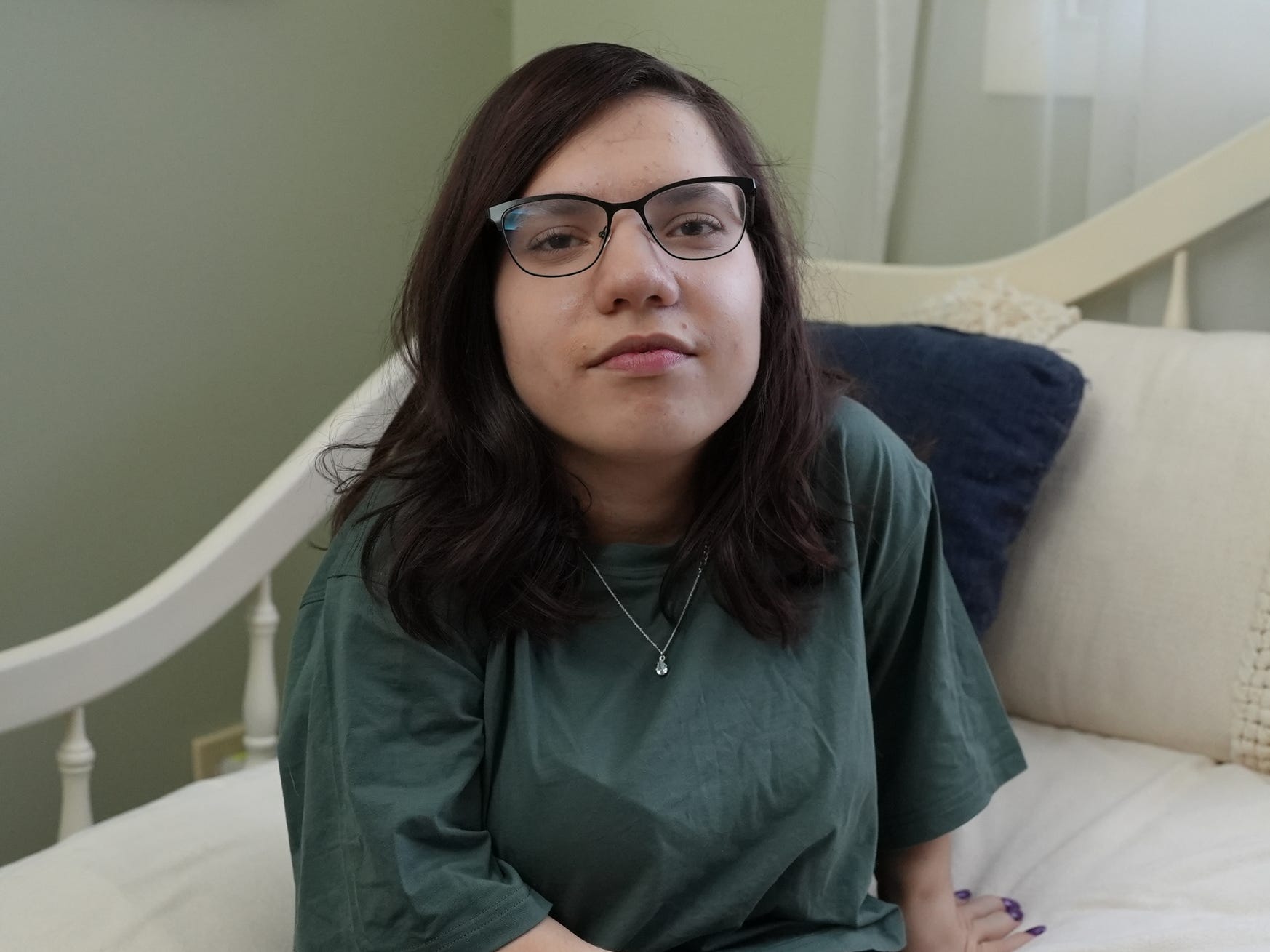 natalia grace mans sitting on her bed, smiling slightly and wearing glasses. she's wearing a green t-shirt, her hair is cut to her collarbone, and her nails are painted. the bedspread is white, with blue and white pillows and a small green stuffed cat toy