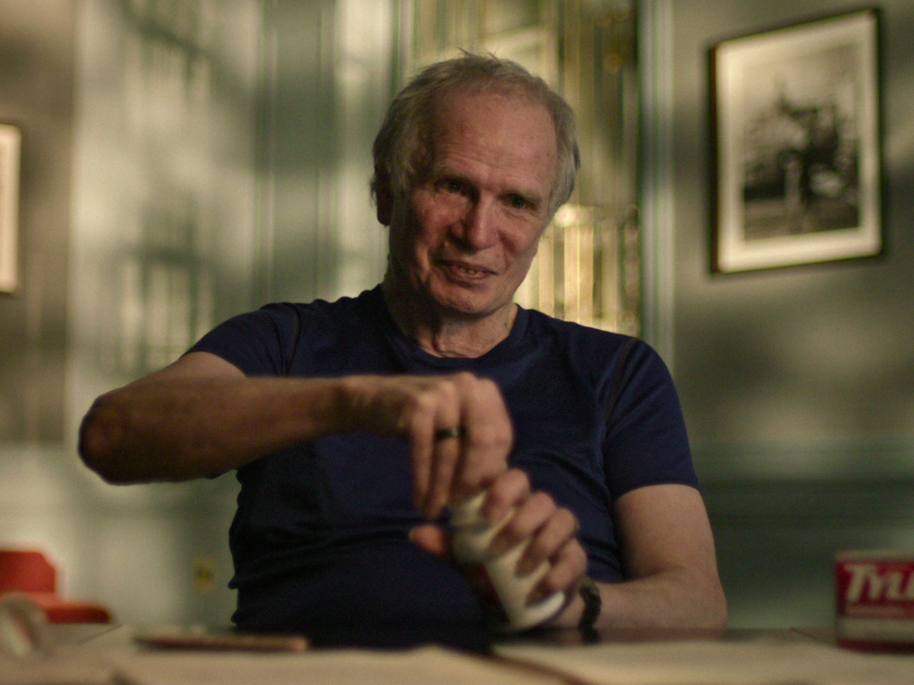 An elderly man with gray hair tries to open a bottle of Tylenol. He's wearing a dark blue T-shirt.