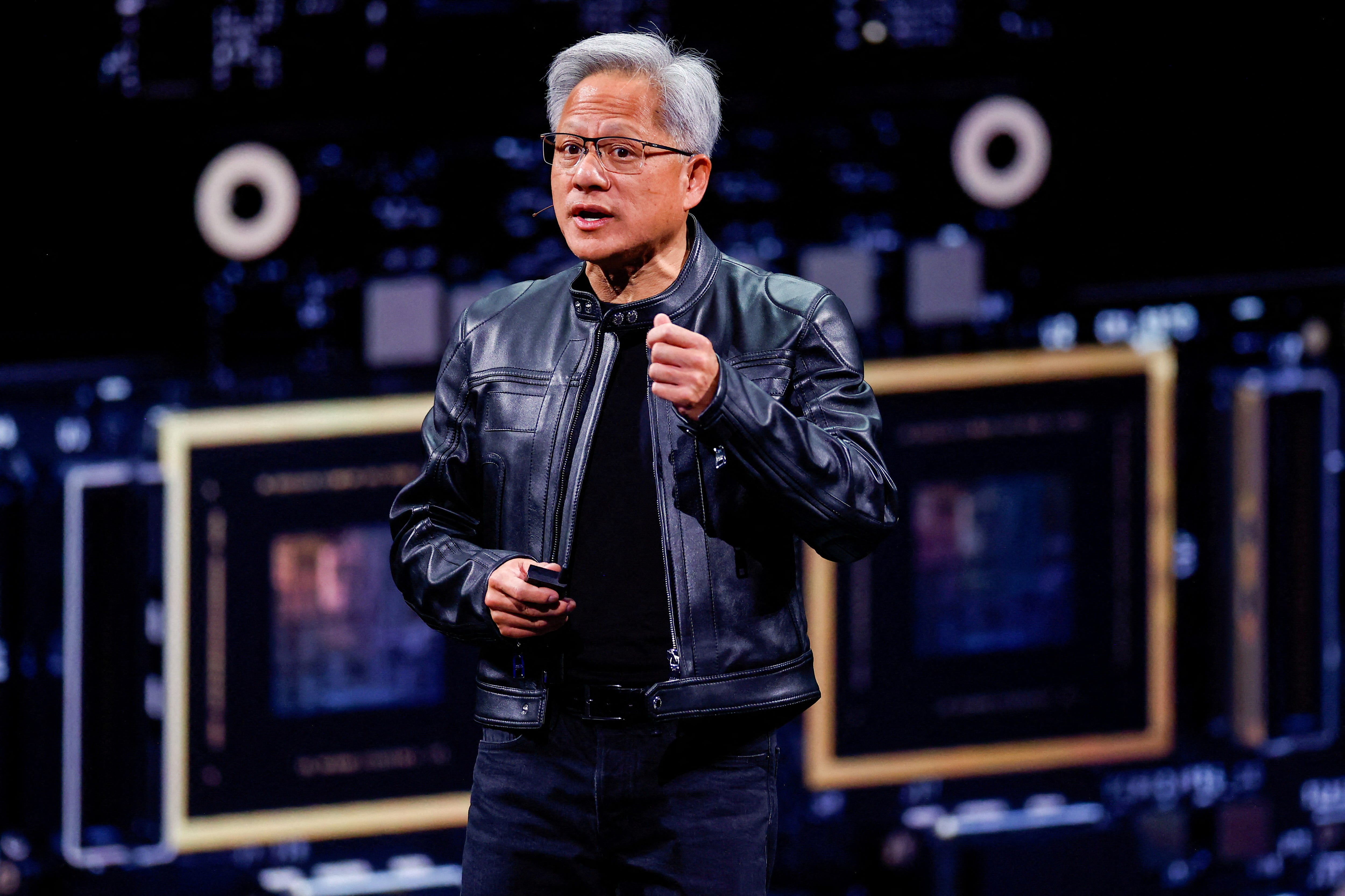 A man (Jensen Huang) wearing all black and a black leather jacket speaks on a stage in front of an enlarged image of a graphics processing unit.