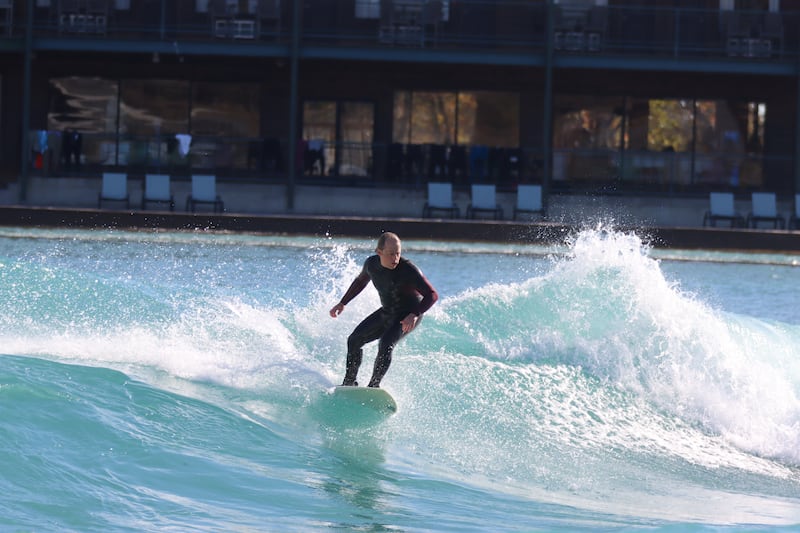 A man in a dry suit surfing on a surf board, in front of a resort-style building