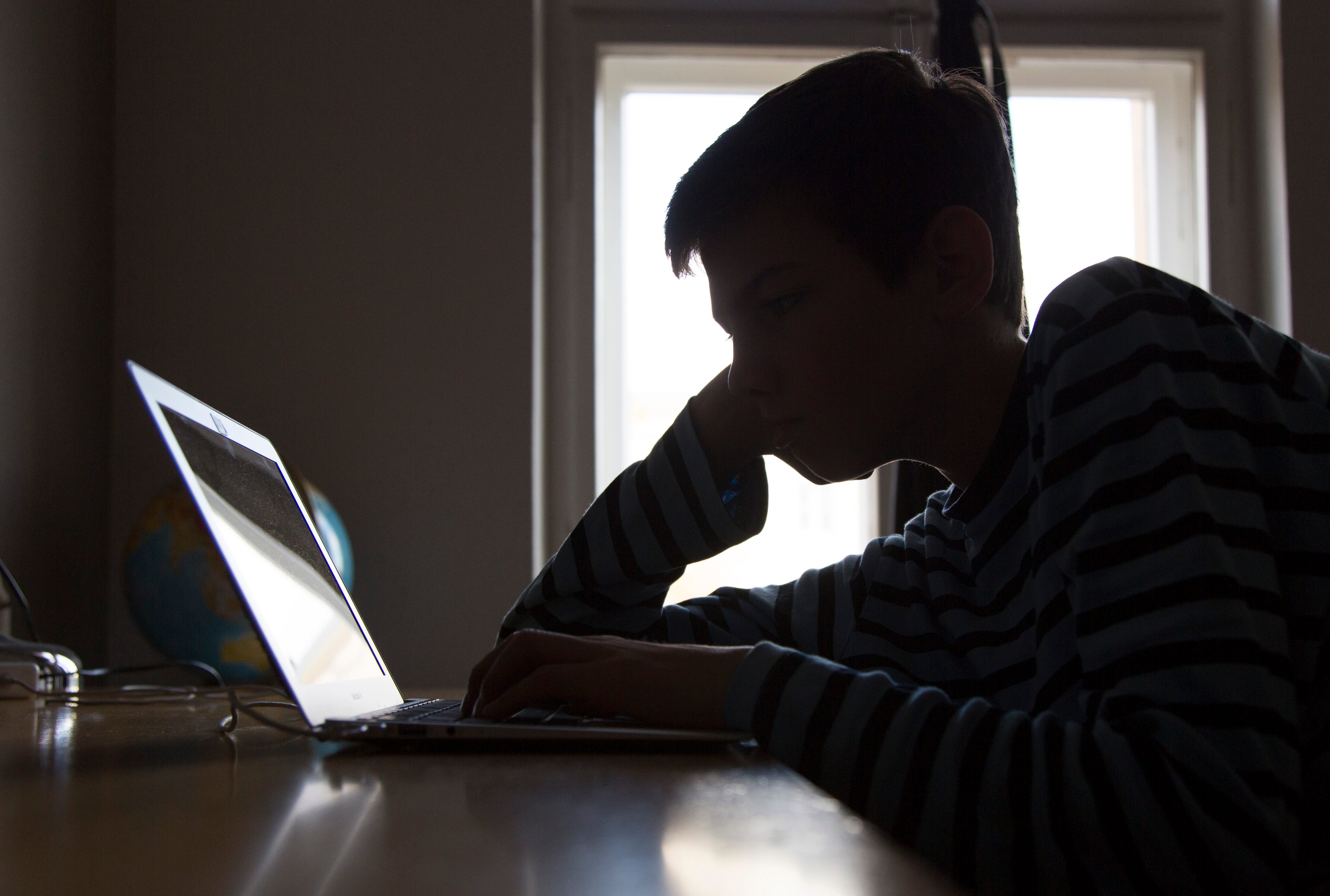 A staged photo of a boy sitting in shadow at a laptop computer.