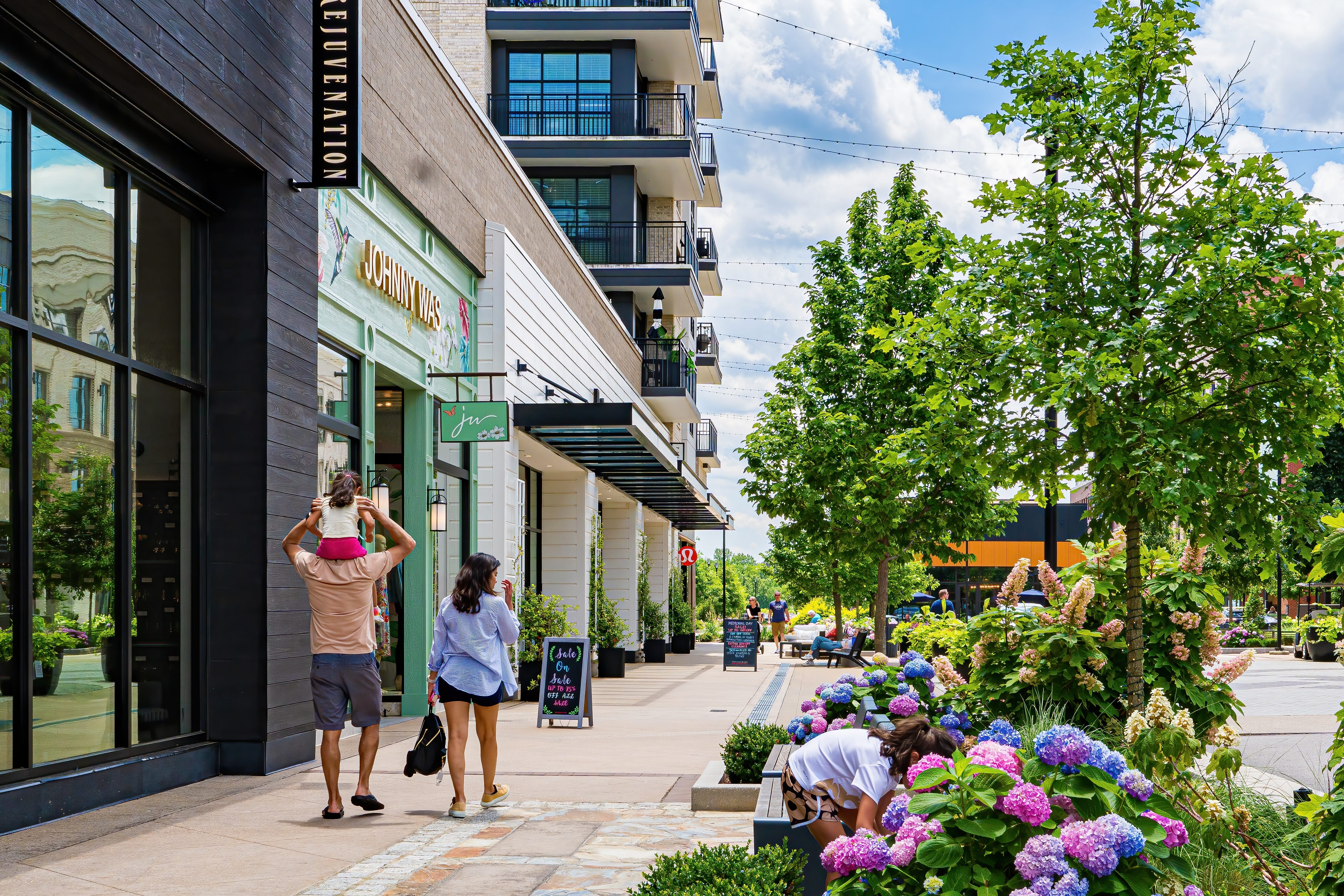 A family walks down a sidewalk lined with stores that has pink and purple hydrangeas and trees planted alongside the road