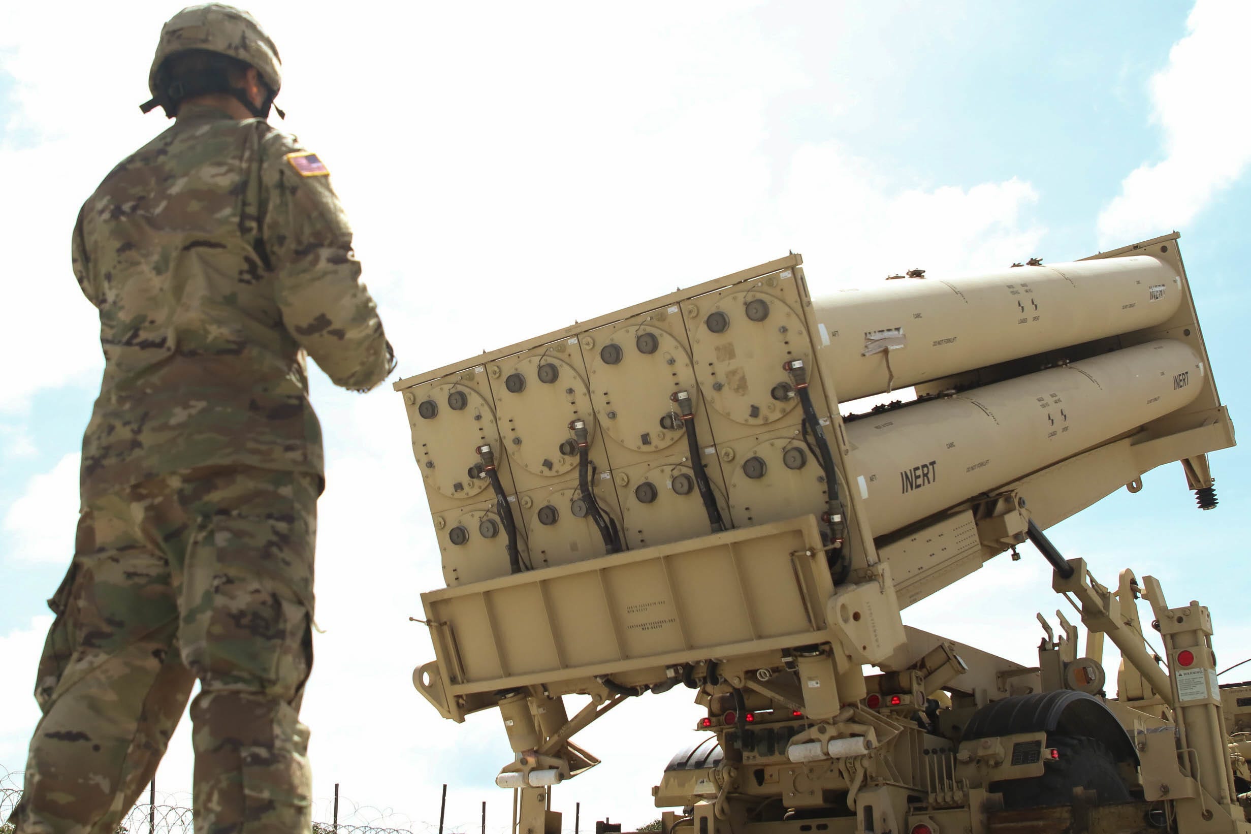 The back of a person in camouflage is seen as they stand behind a missile pallet sitting on a truck. The sky is cloudy and blue.