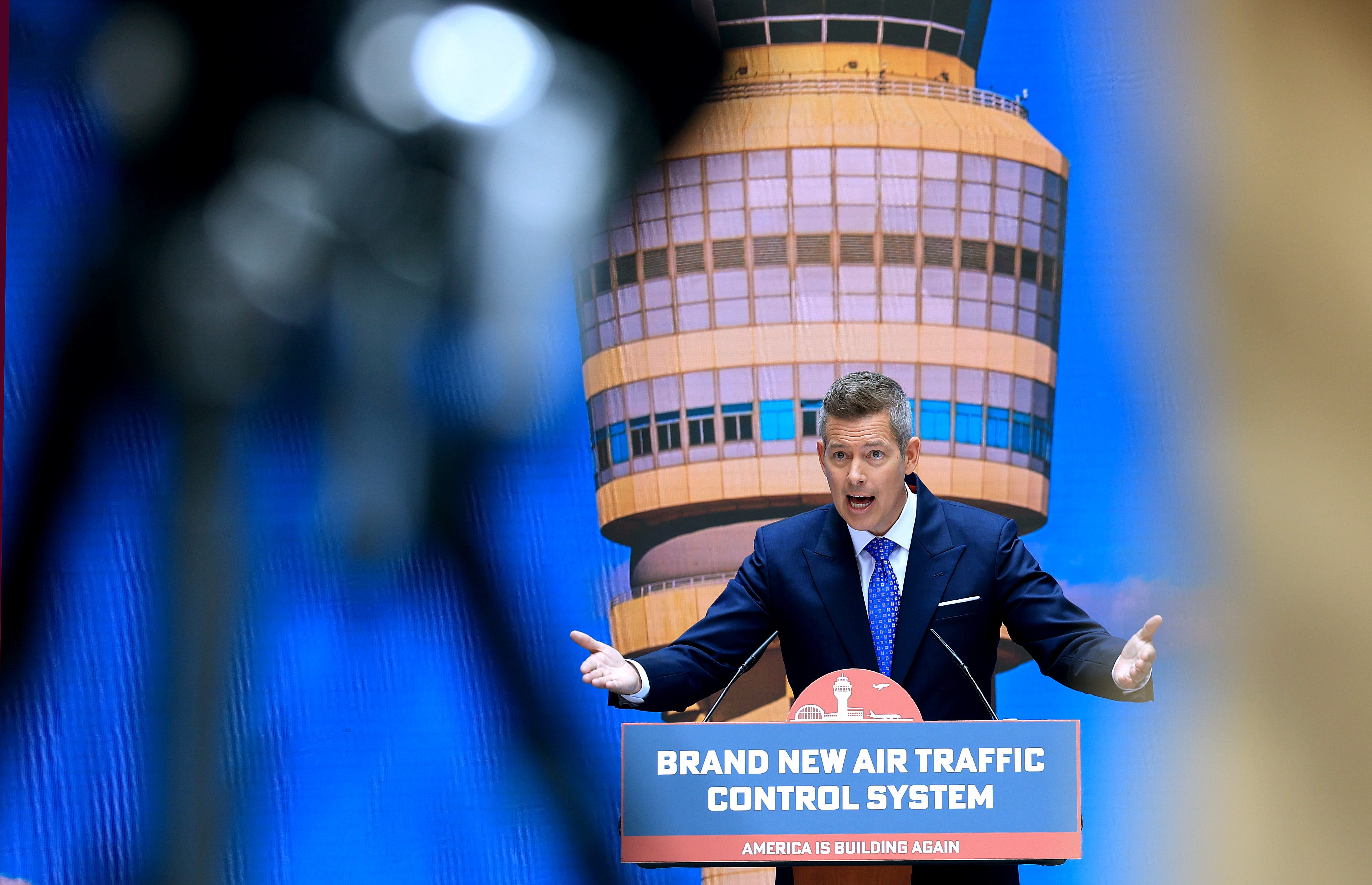 Transportation Secretary Sean Duffy speaks at a press conference behind a podium with an air traffic control tower pictured behind him.