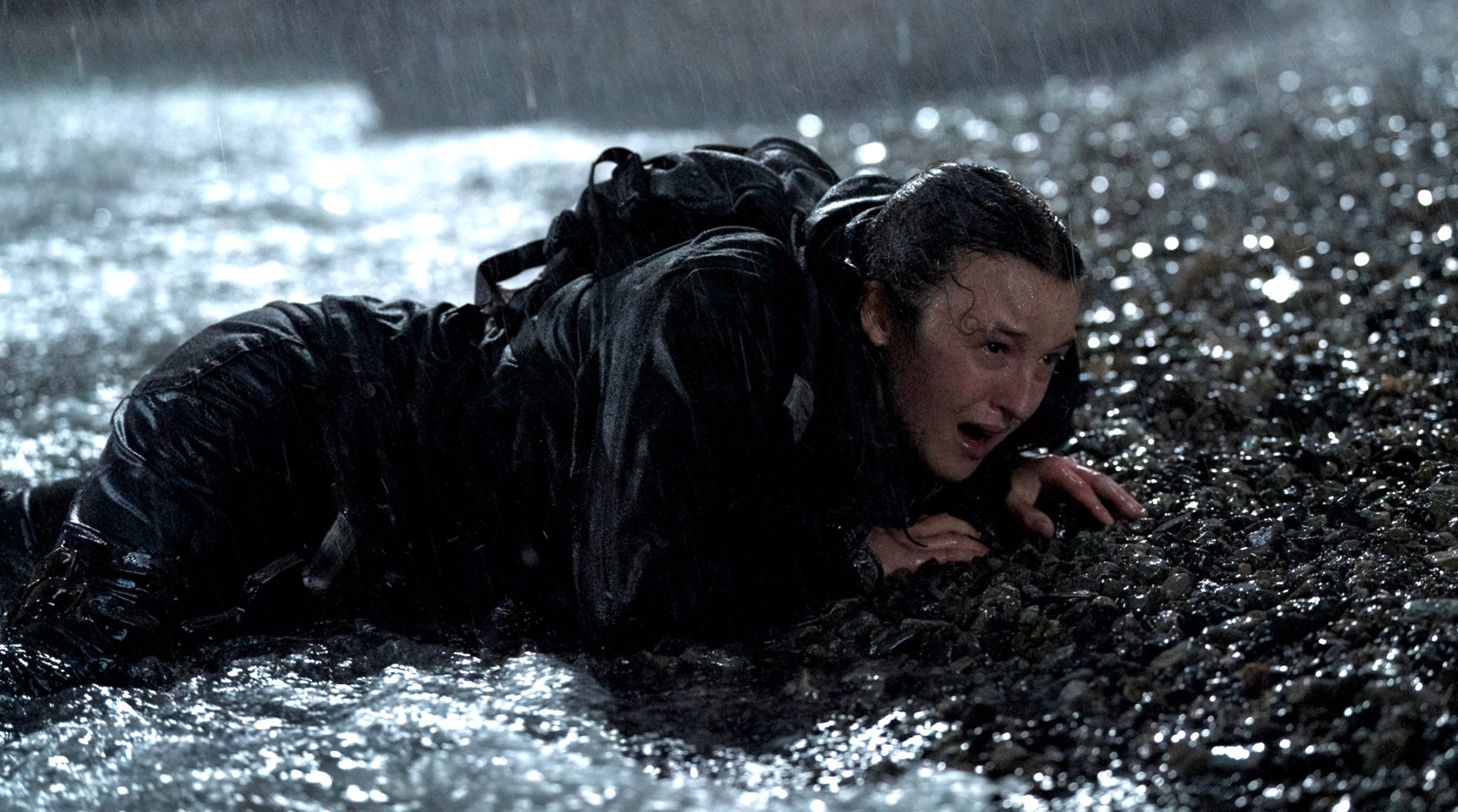 A young woman with long brown hair dressed in dark clothes crawls out of the sea onto the shore. She is completely soaked by the sea and the heavy rain.