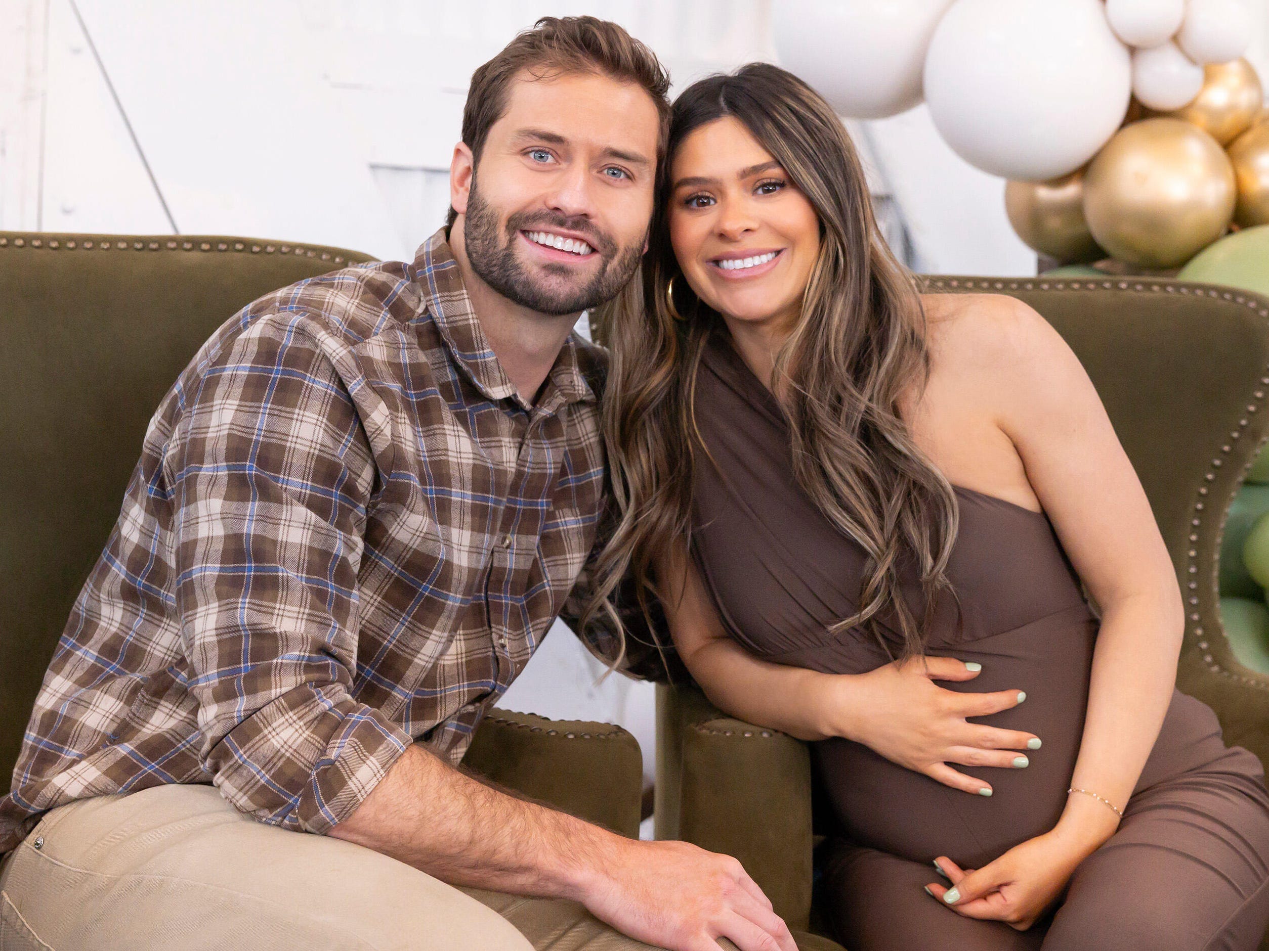 A still from "The Secret Lives of Mormon Wives" featuring Dakota Mortensen in a plaid brown shirt and brown pants hugging his partner Taylor Frankie Paul, who is in a brown dress, while they sit on two green chairs.