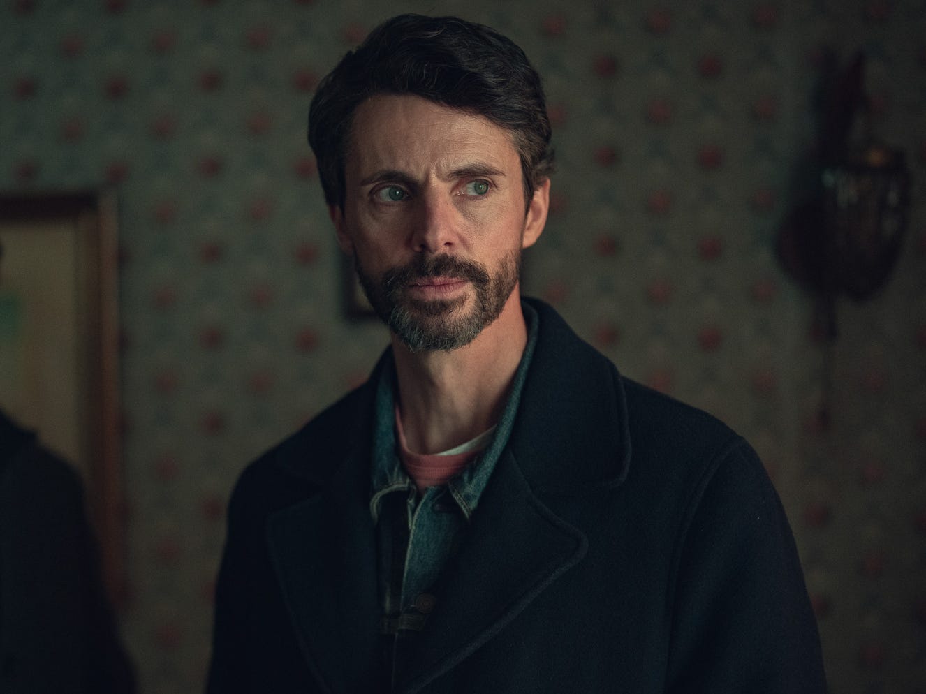A man stood inside a living room. He has neat brown hair with a short brown beard. He's wearing a black coat with a denim shirt underneath.