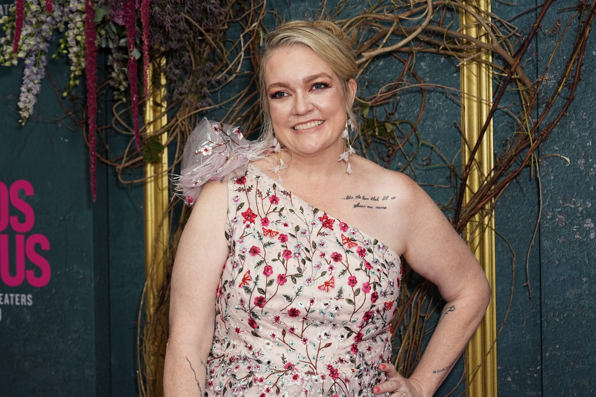 Colleen Hoover smiles in front of a blue background with greenery.