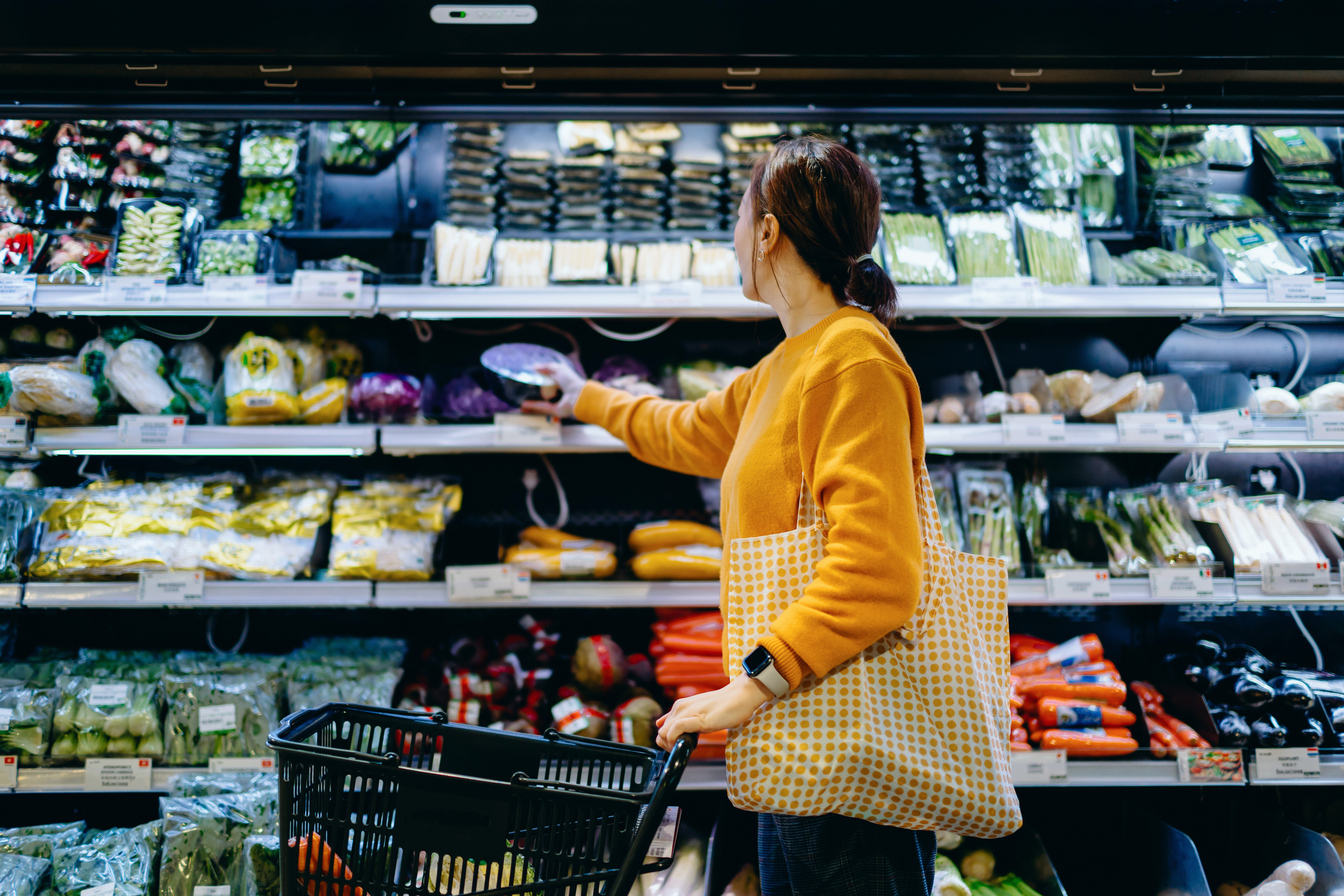 A woman pushes a shopping cart and pulls items off a shelf in a grocery store.