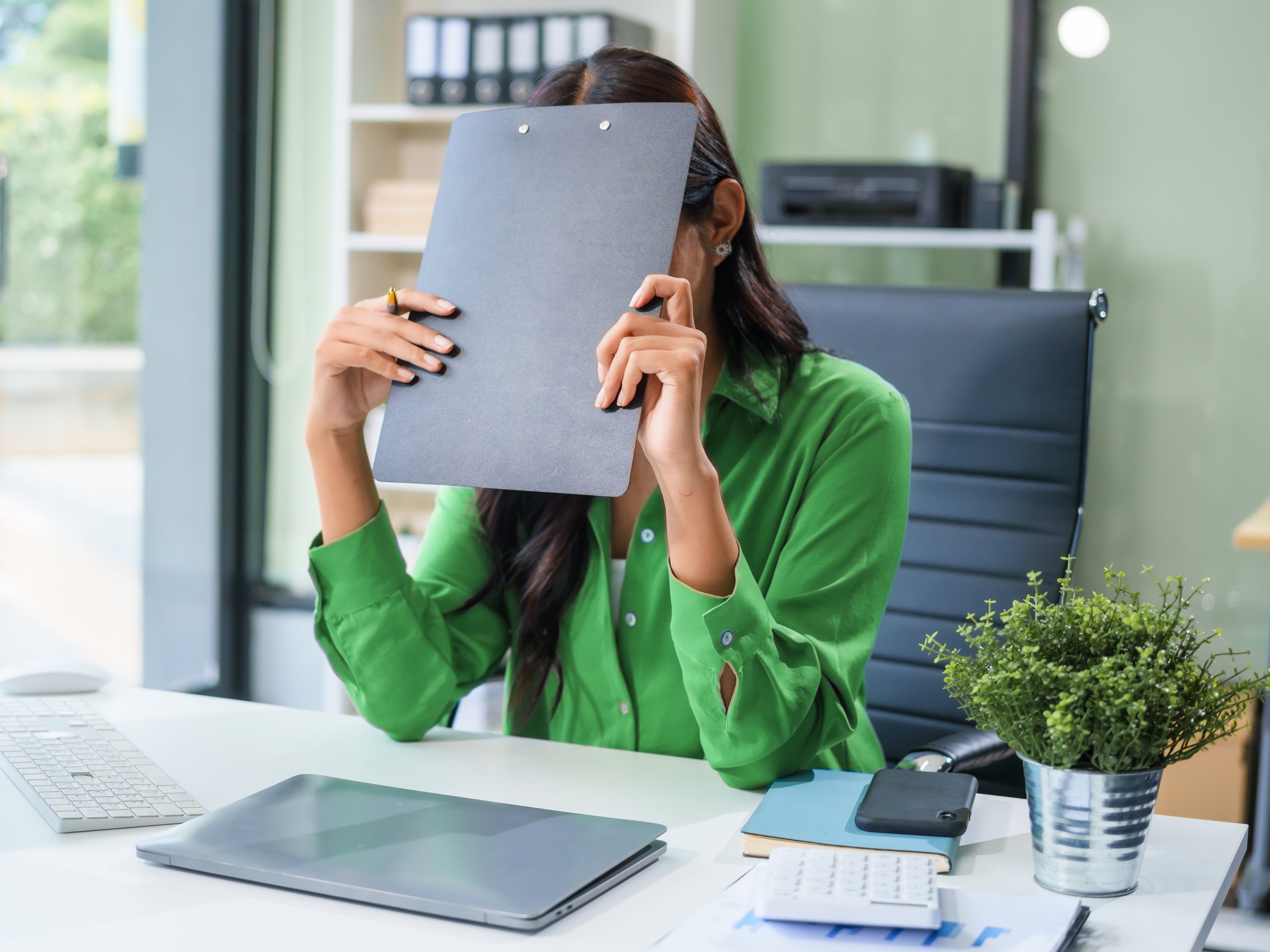 A woman wearing a green shirt sits in a corporate office, olding a folder in front of her face.