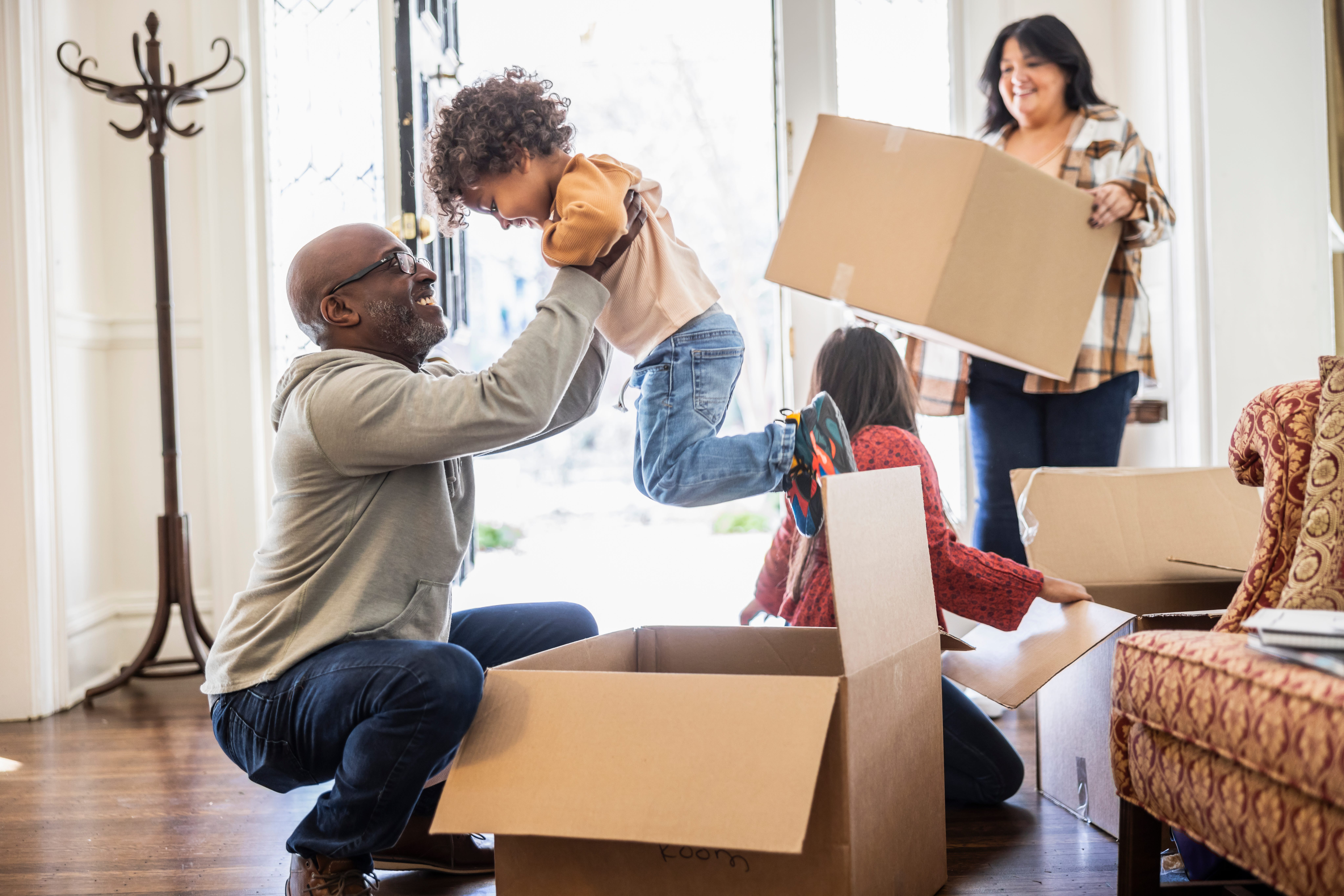 A man lifting up a small child over some boxes, another child and a woman to the right with boxes in a house doorway.