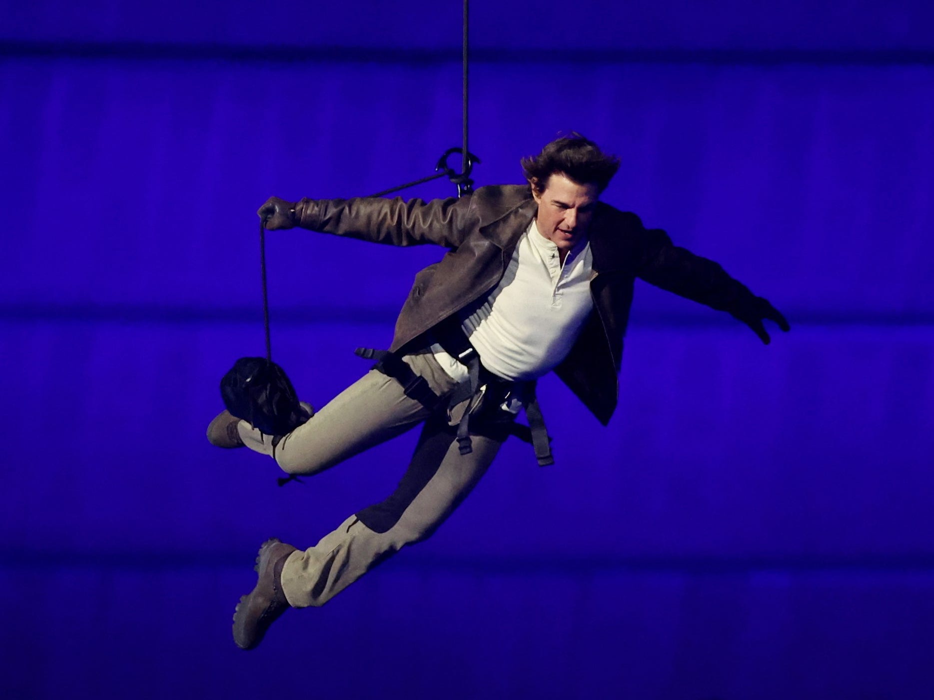 A man in a brown leather jacket, a white shirt, and beige trousers falling through the air in a harness against a blue background.