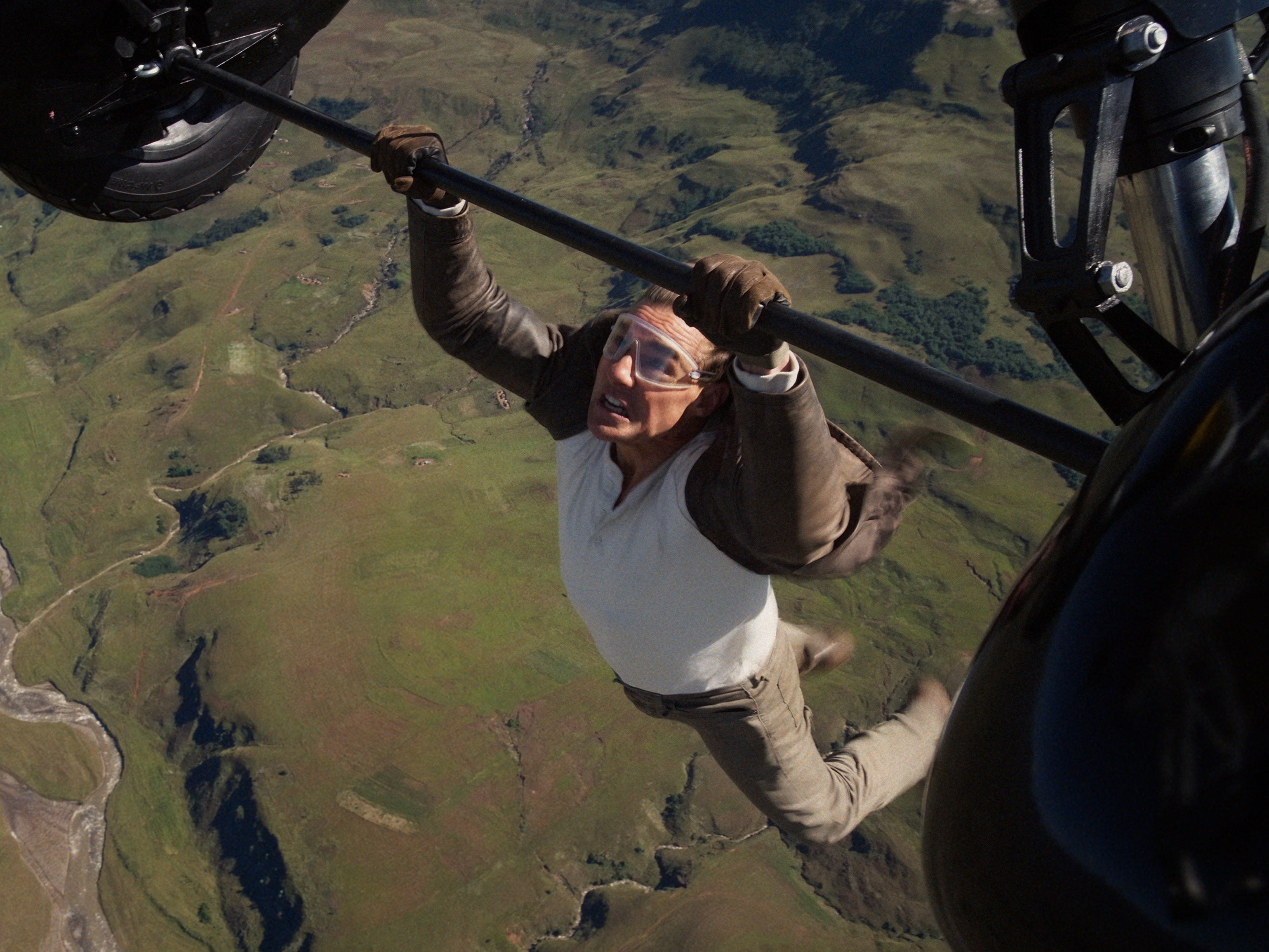 A man wearing a brown leather jacket. a white long-sleeved shirt, brown pants, and shoes. He's clinging onto a set of aircraft wheels while flying without a harness. He's also wearing a pair of goggles.