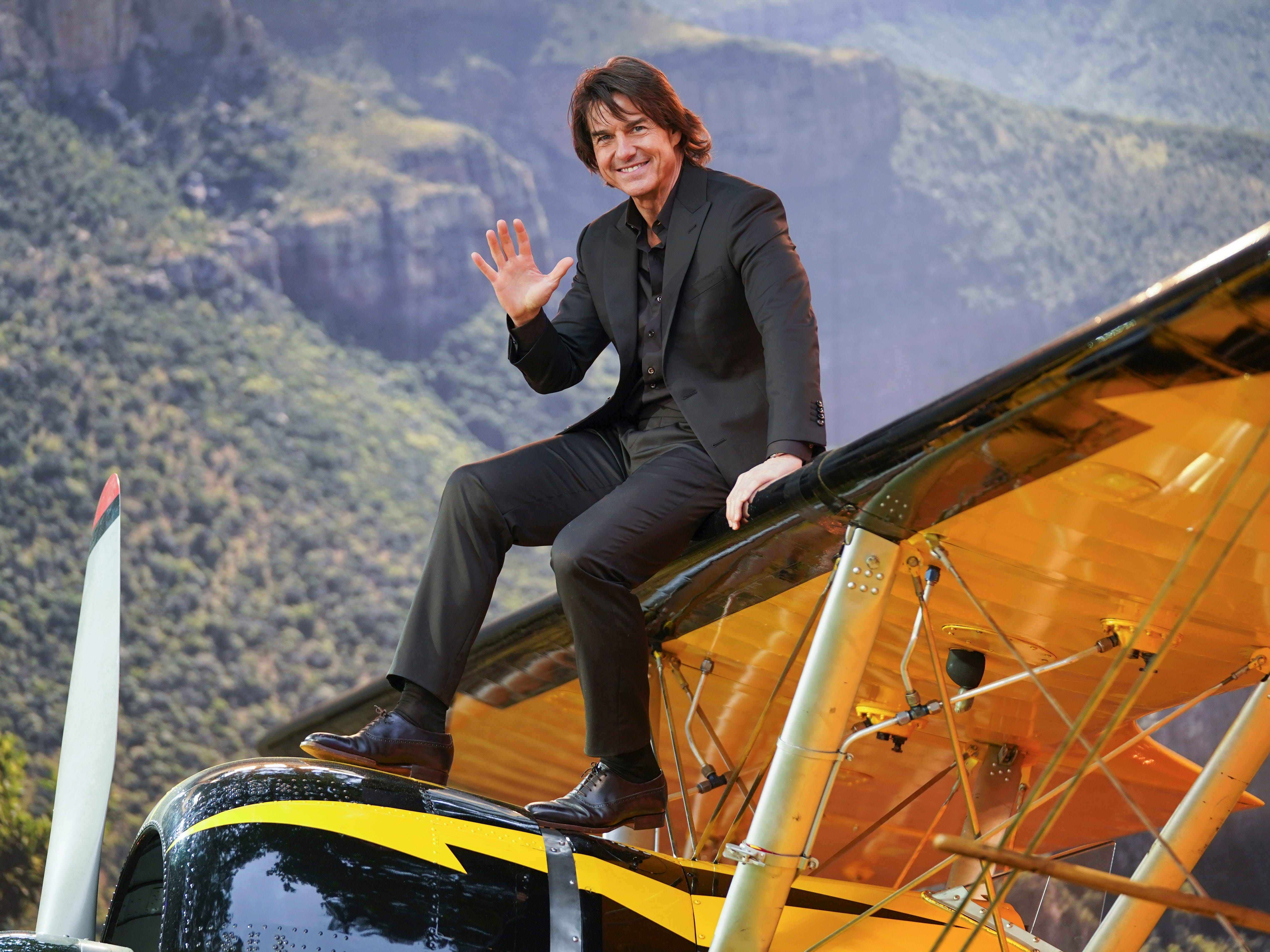 A man with long brown hair is dressed in an all black suit with an open collar. He's sitting on a yellow bi-plane.