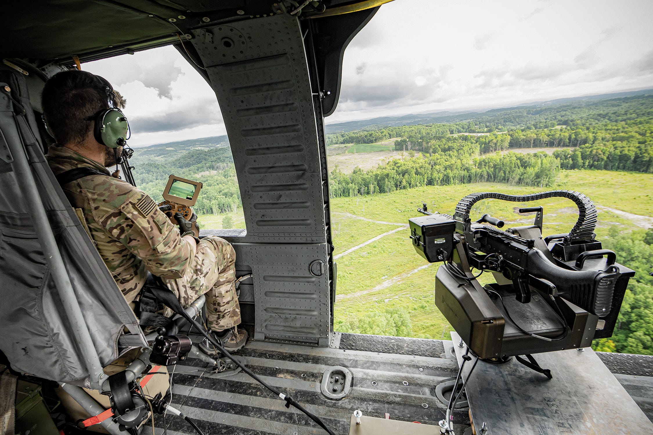 An automated machine gun sits in a helicopter with a man remote-controlling it. Outside of the helicopter is a green field and forest with an overcast sky.